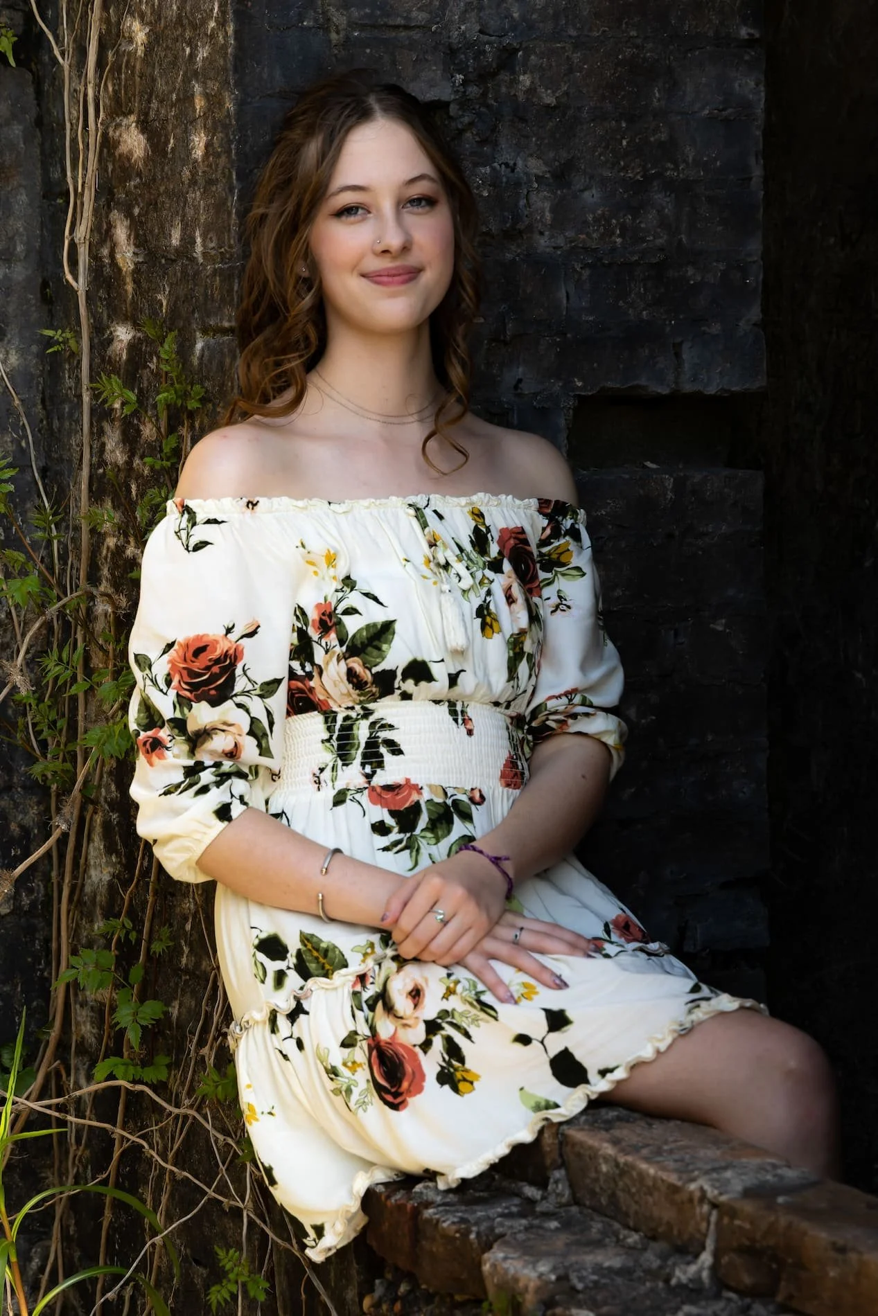 Senior Session at Fort Pickens, FL A young woman with wavy brown hair and a nose piercing sitting on a brick ledge near a dark stone wall and green foliage, wearing an off-the-shoulder white floral dress.
