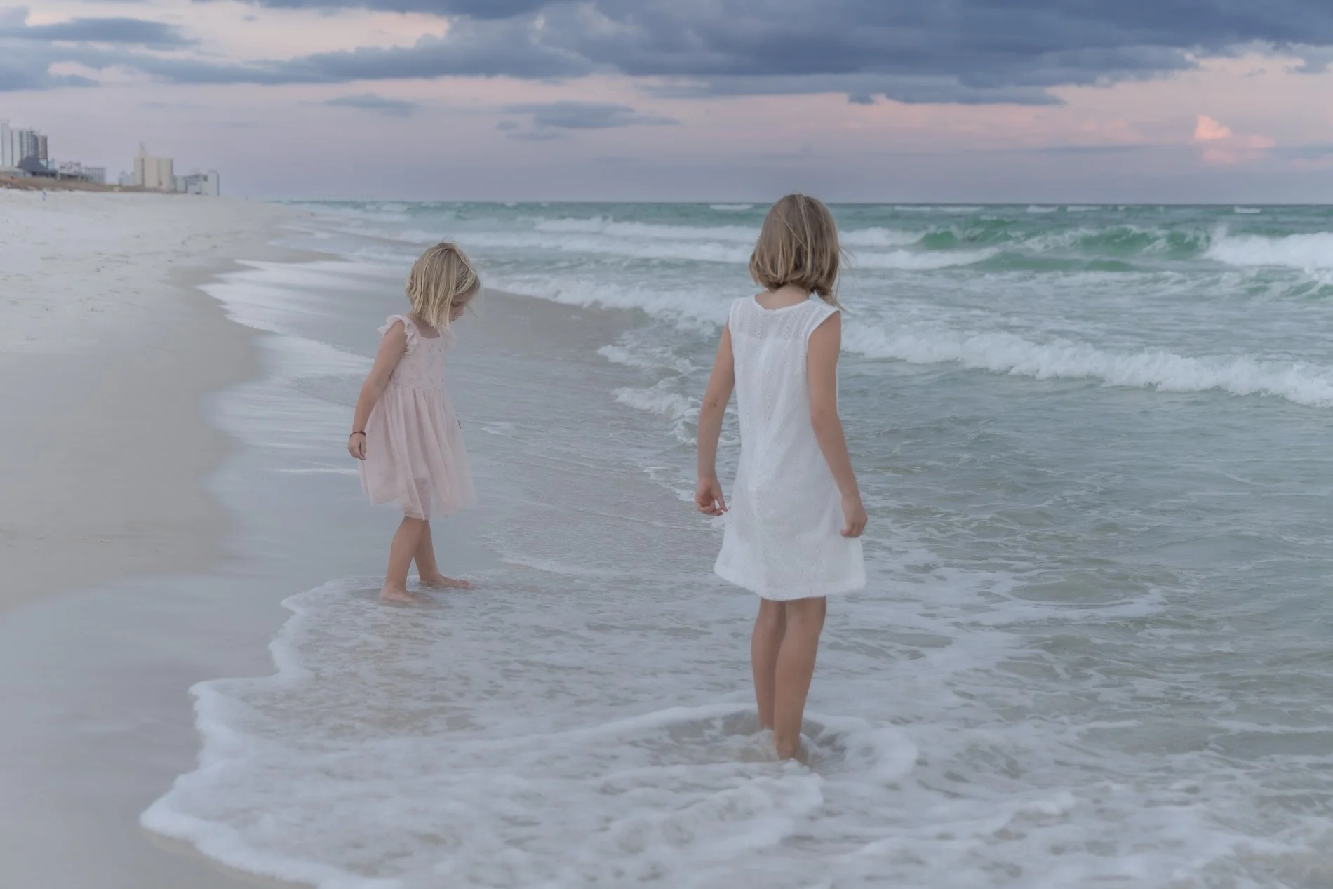 Golden hour family portrait at Pensacola Beach Two young girls in white dresses standing at the edge of the ocean on a beach, looking at the water as waves gently crash around their feet.