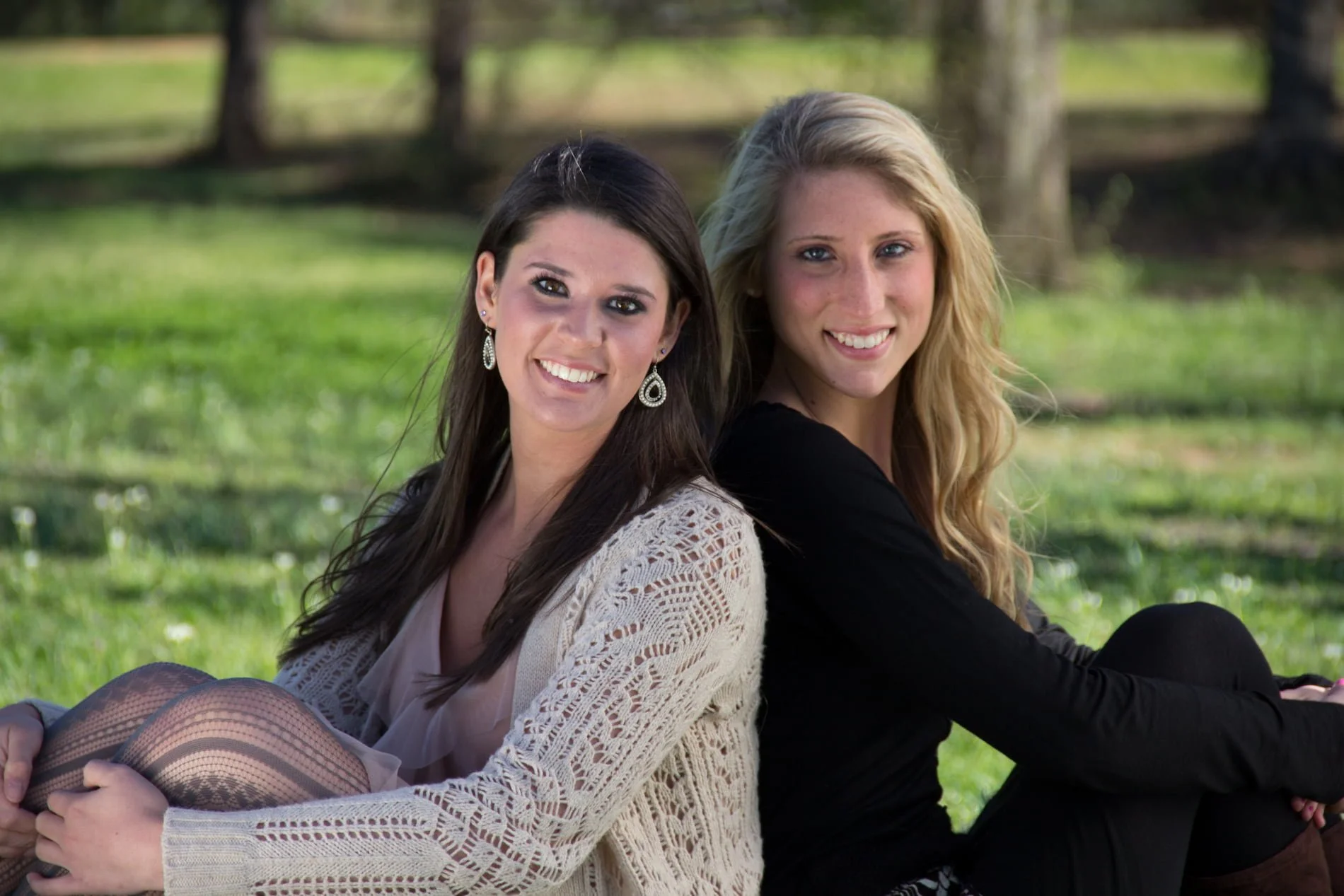 Two young, graduating senior women friends sitting on grass in a park, smiling at the camera, with trees in the background.