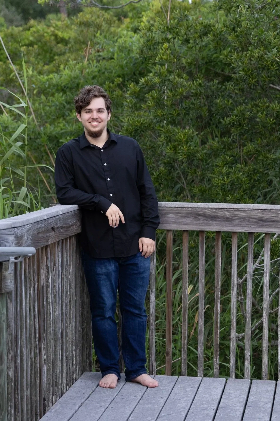 Gulf Breeze senior portraits — young man in black shirt and jeans standing barefoot on a wooden boardwalk, Gulf Coast natural light