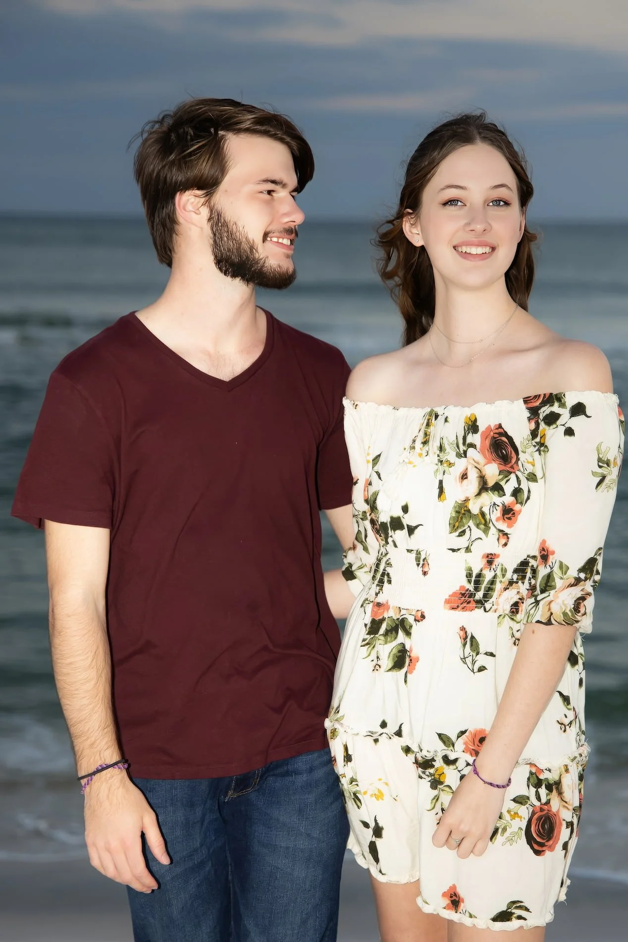 Blue hour Pensacola Beach Portrait A young couple standing on the beach, with the ocean and sky in the background. The man has brown hair and a beard, wearing a maroon T-shirt and jeans. The woman has long brown hair and is wearing an off-shoulder fl