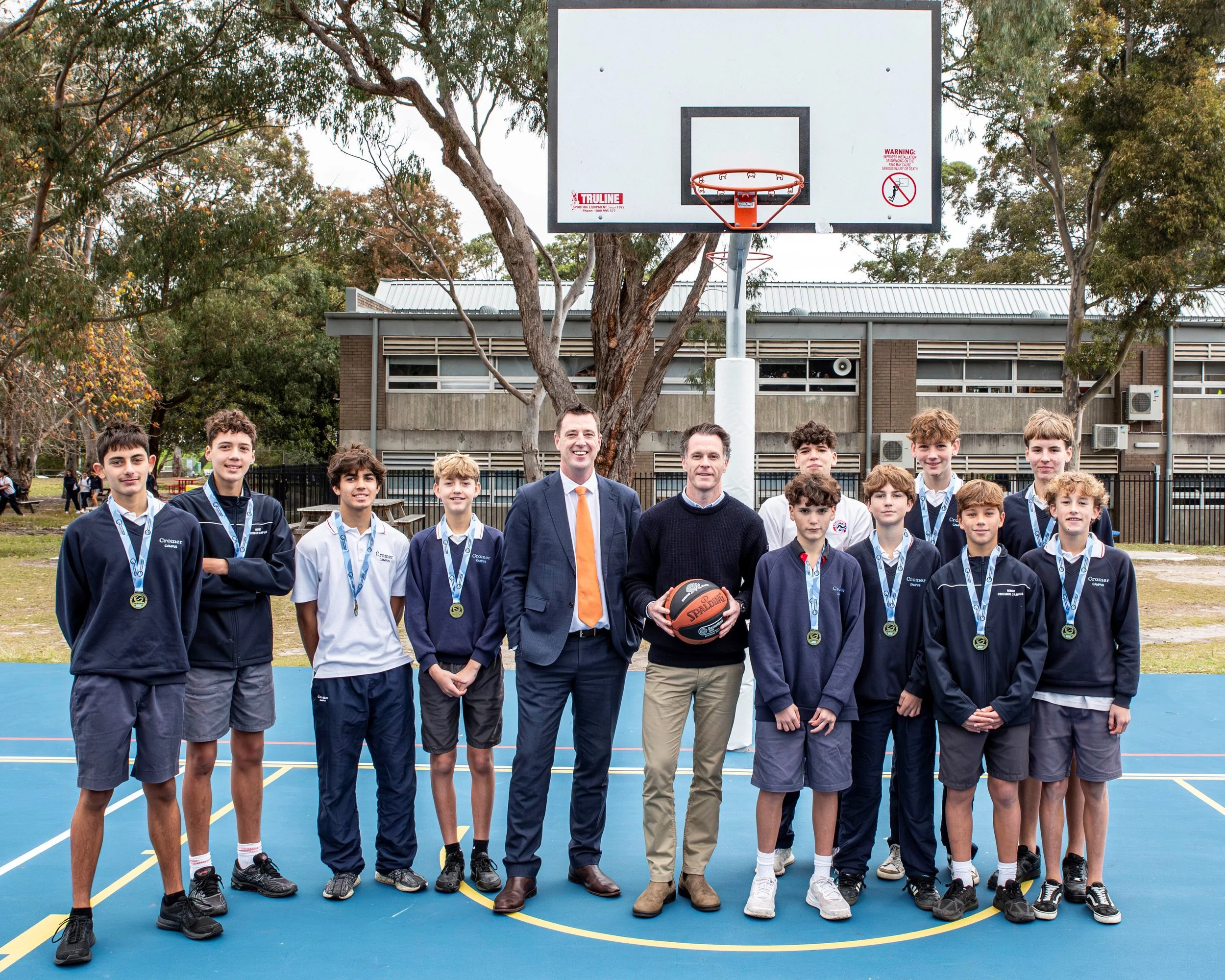 School basketball team posing with coaches on an outdoor basketball court, with medals, a basketball hoop, and a basketball.