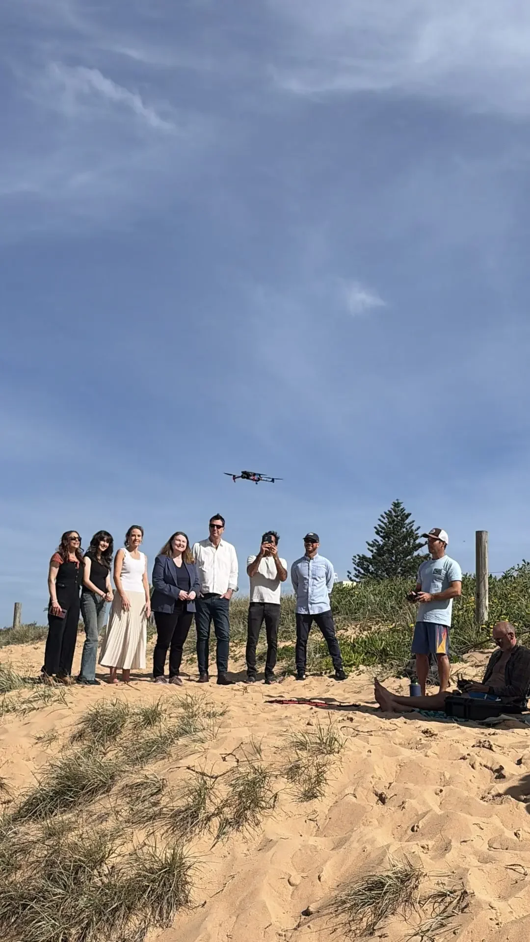 Group of people standing on a sandy beach dune beneath a hovering drone