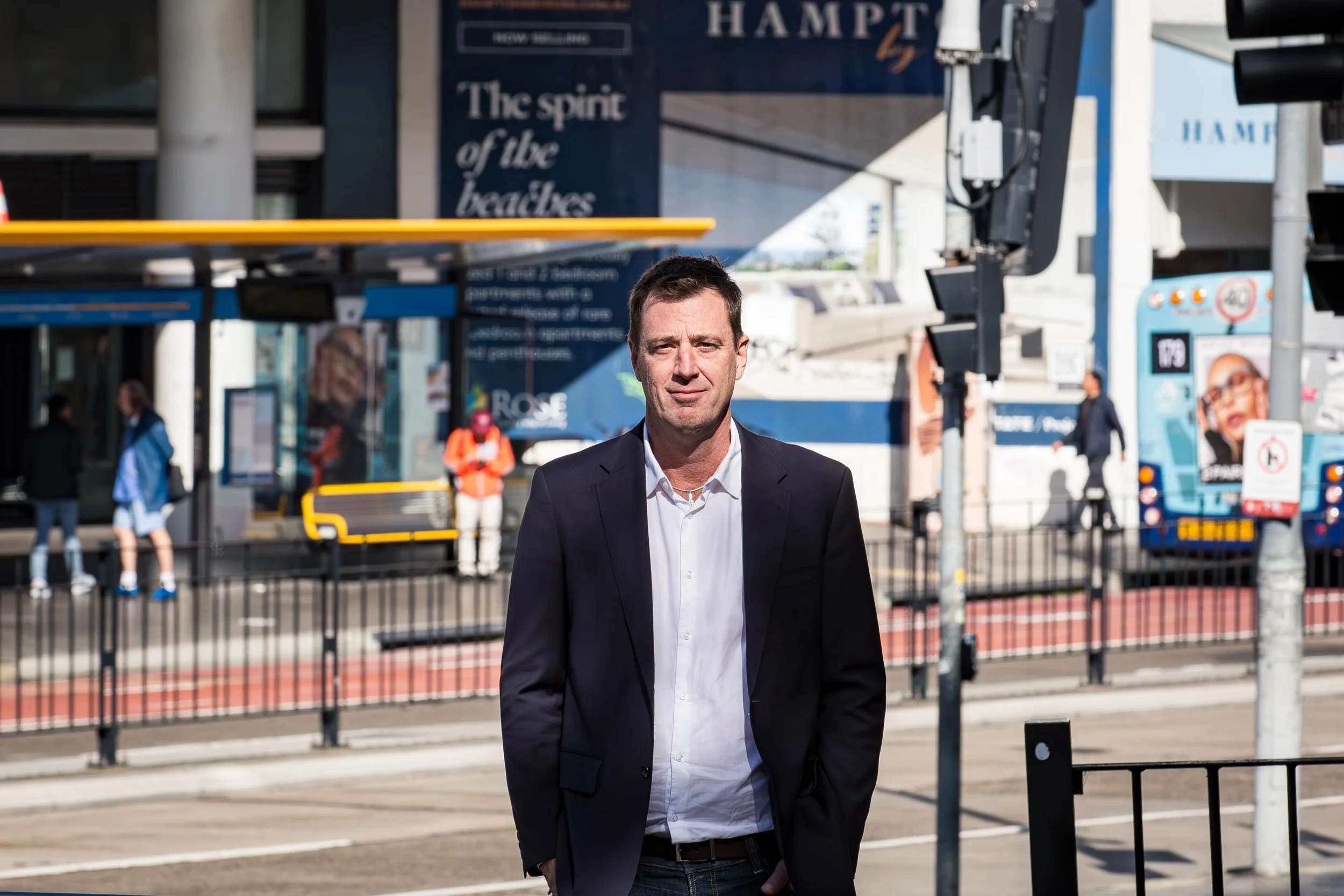 Man in a dark blazer standing outside a Hampton by Hilton hotel near a city bus stop