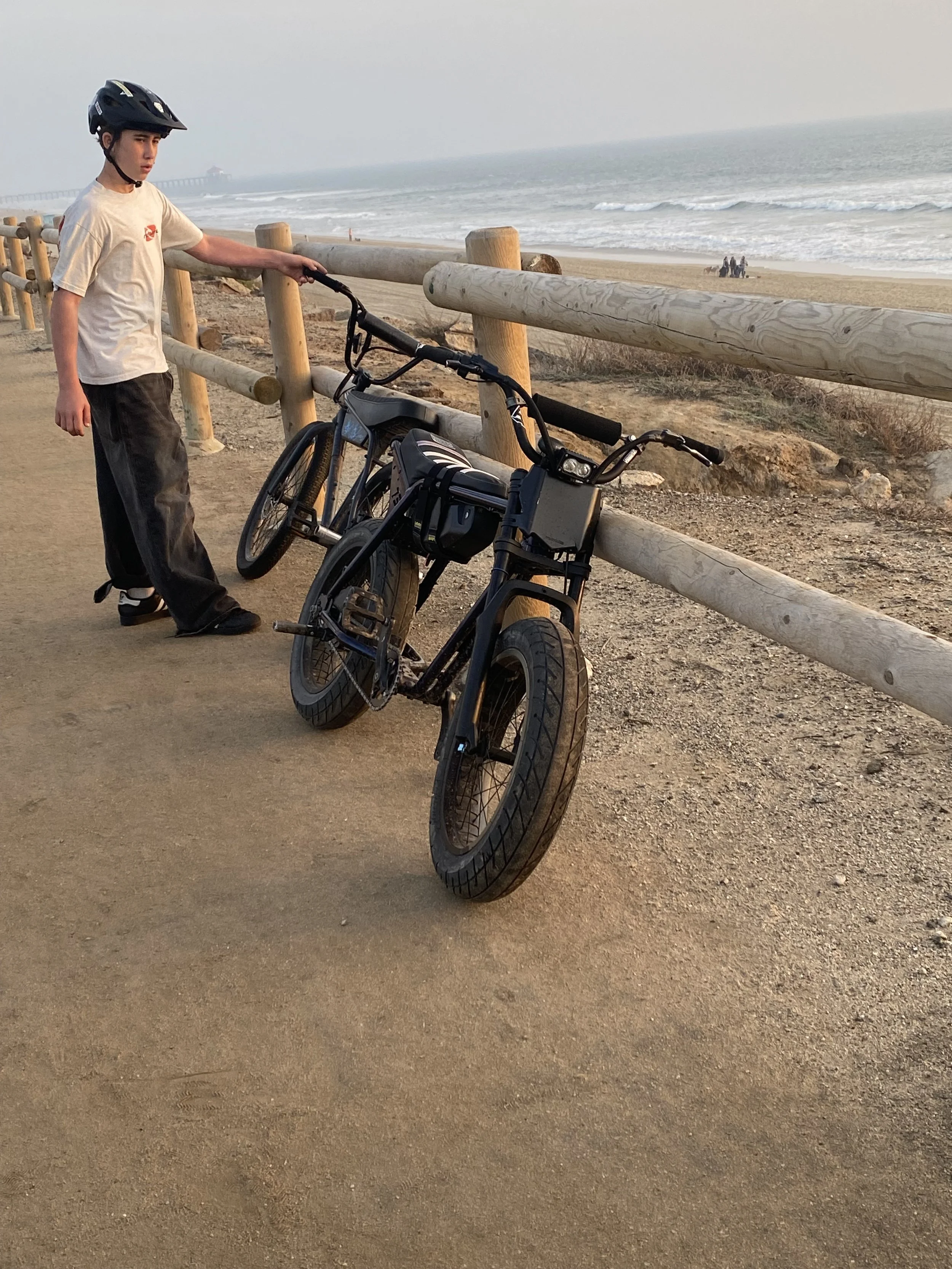A boy wearing a helmet and a white T-shirt standing next to two black electric bikes on a sandy path by the beach, with the ocean and a distant pier in the background.