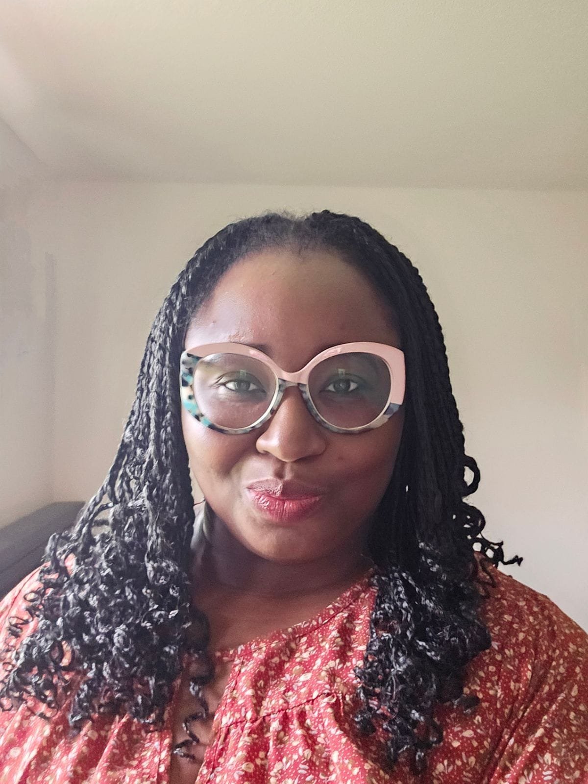 A woman with dark skin wearing large, round glasses with a colorful pattern on the frame. She has long, curly black hair and is wearing a red patterned blouse. She is smiling slightly indoors with beige walls behind her.
