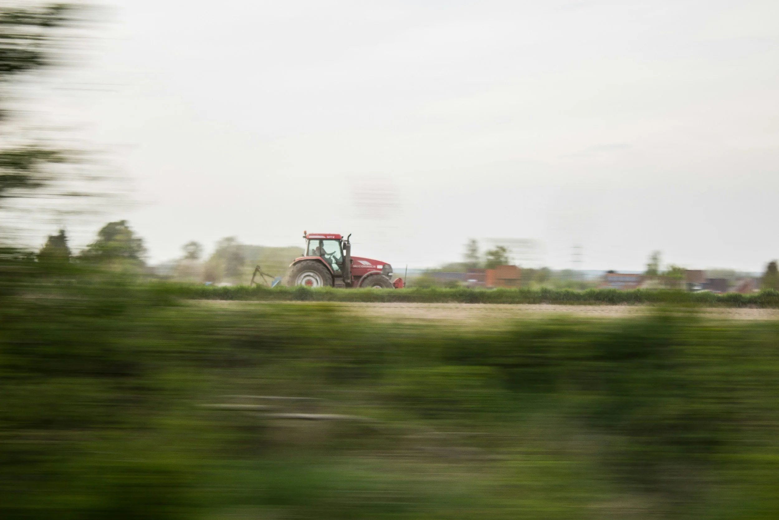 A red tractor working on a farm field during daytime, with a background of trees and buildings, viewed from a moving vehicle.