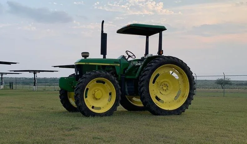 Green and yellow tractor on a grassy field with a cloudy sky and solar panels in the background.
