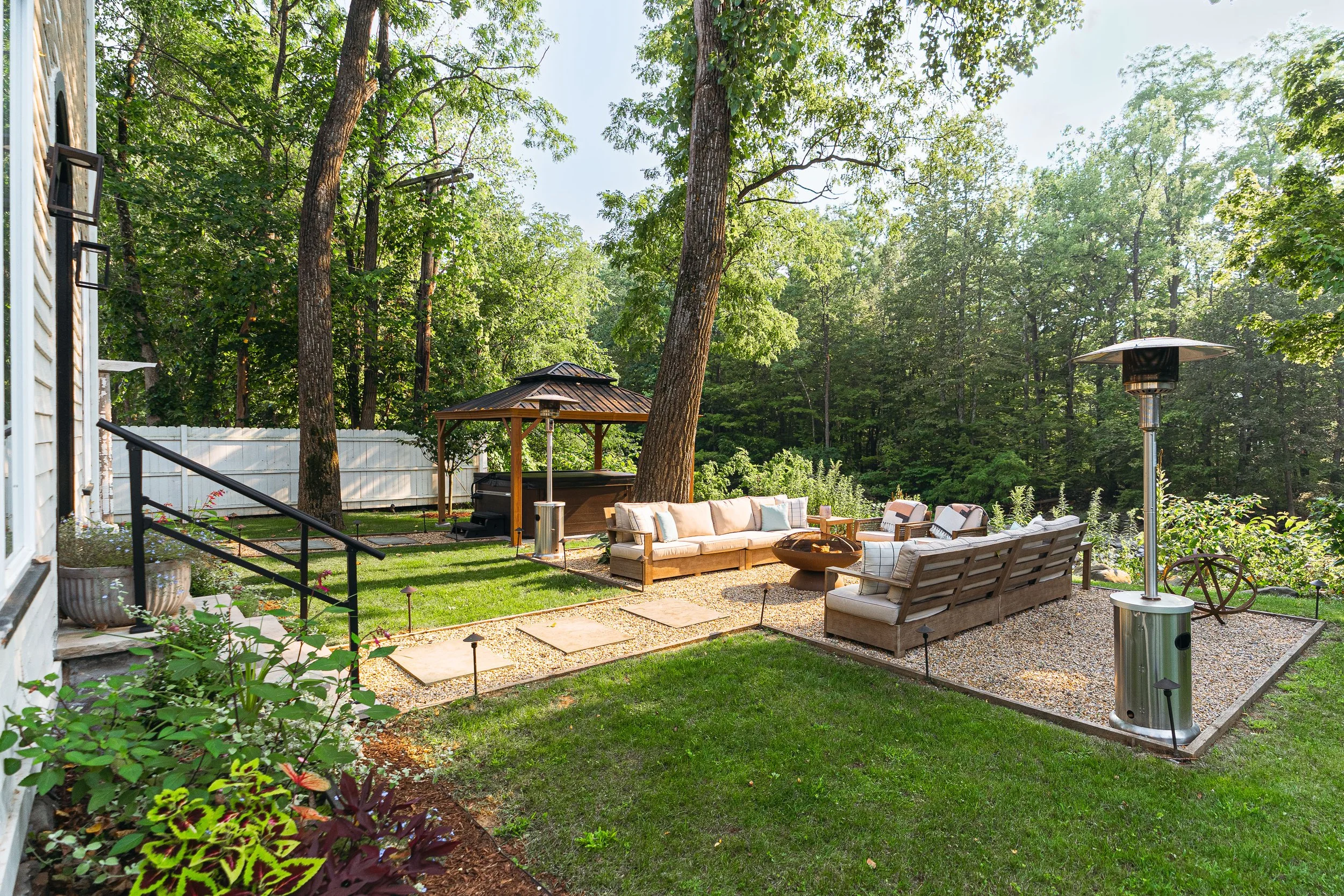 A backyard patio with outdoor furniture, a hot tub under a gazebo, a patio heater, and surrounded by trees and greenery.