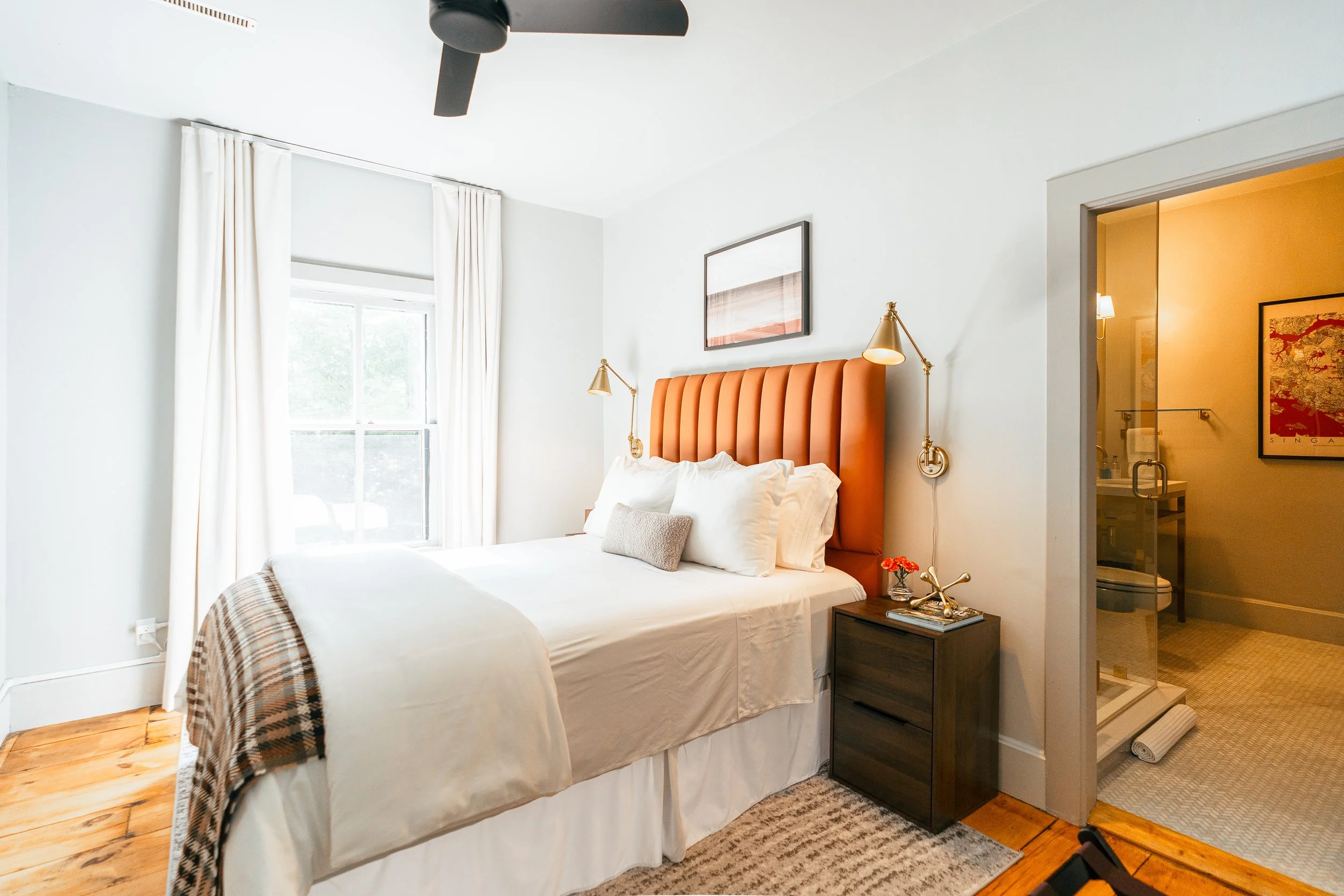 A bedroom with a large bed featuring white bedding and an orange upholstered headboard, bedside lamps, a wooden nightstand with decor, and a window with white curtains. There is an adjoining bathroom with a glass shower, visible through an open doorway.