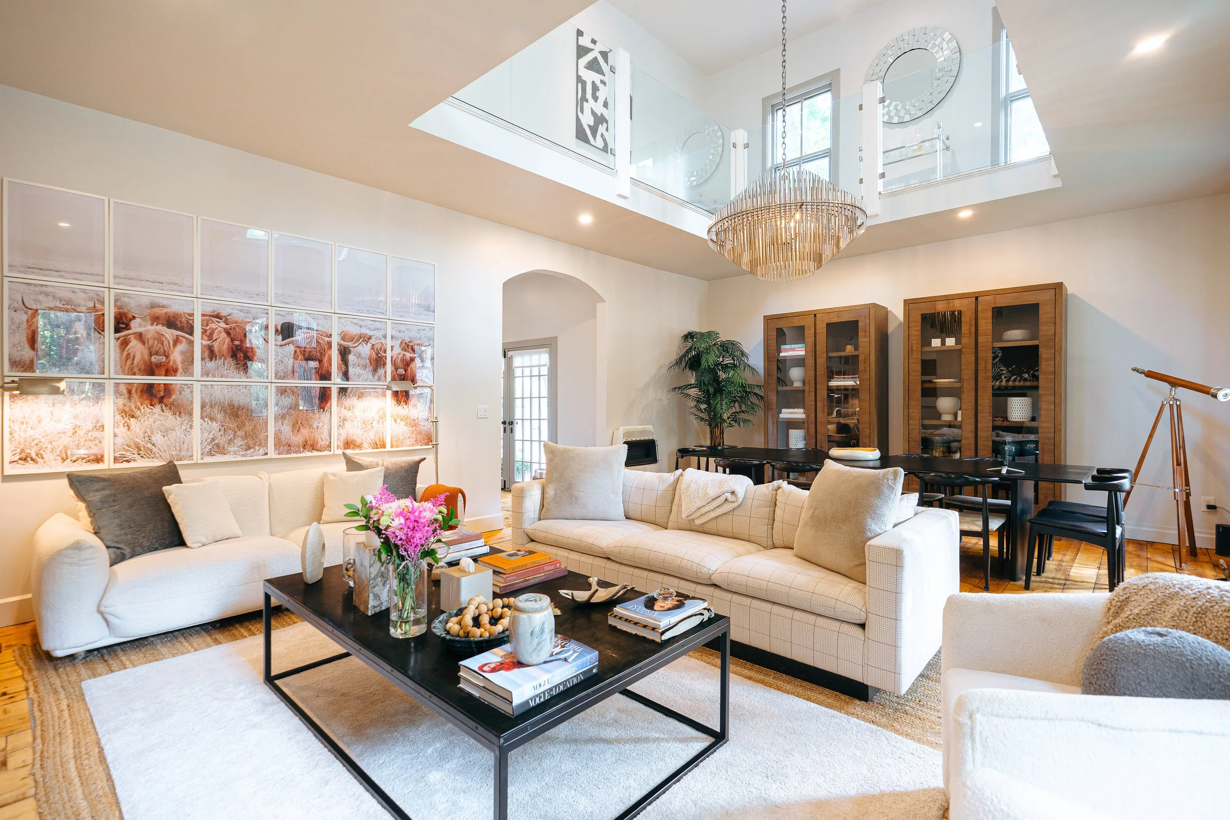 Spacious living room with white sofas, a black coffee table with books and decorations, large wall art of cows, a wooden dining table with black chairs, a tall wooden cabinet, a chandelier, and a telescope near the window.
