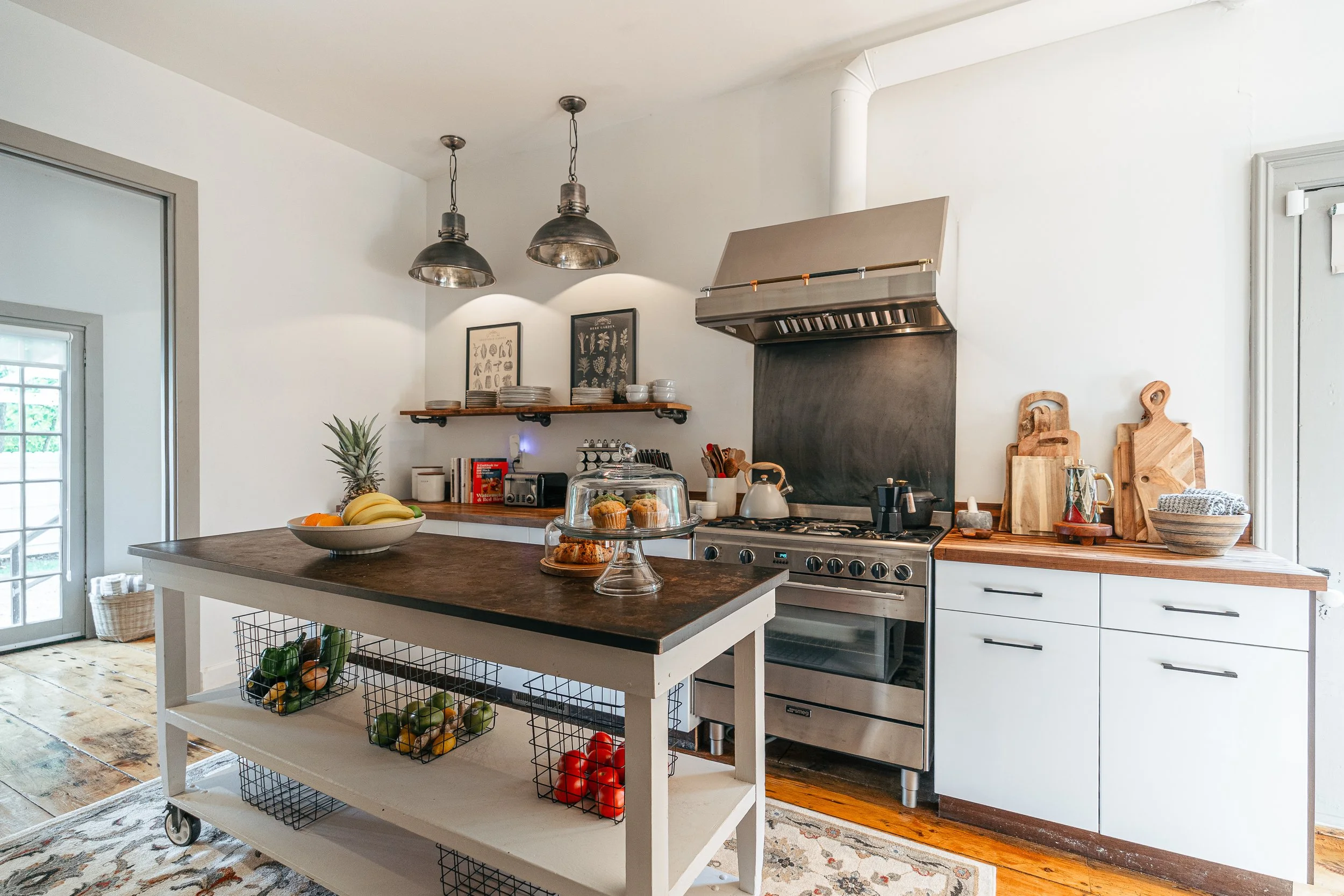Modern kitchen with white cabinets, wooden countertops, stainless steel stove, tropical fruits, baked goods on a cake stand, wooden cutting boards, and a kitchen island with baskets of vegetables.