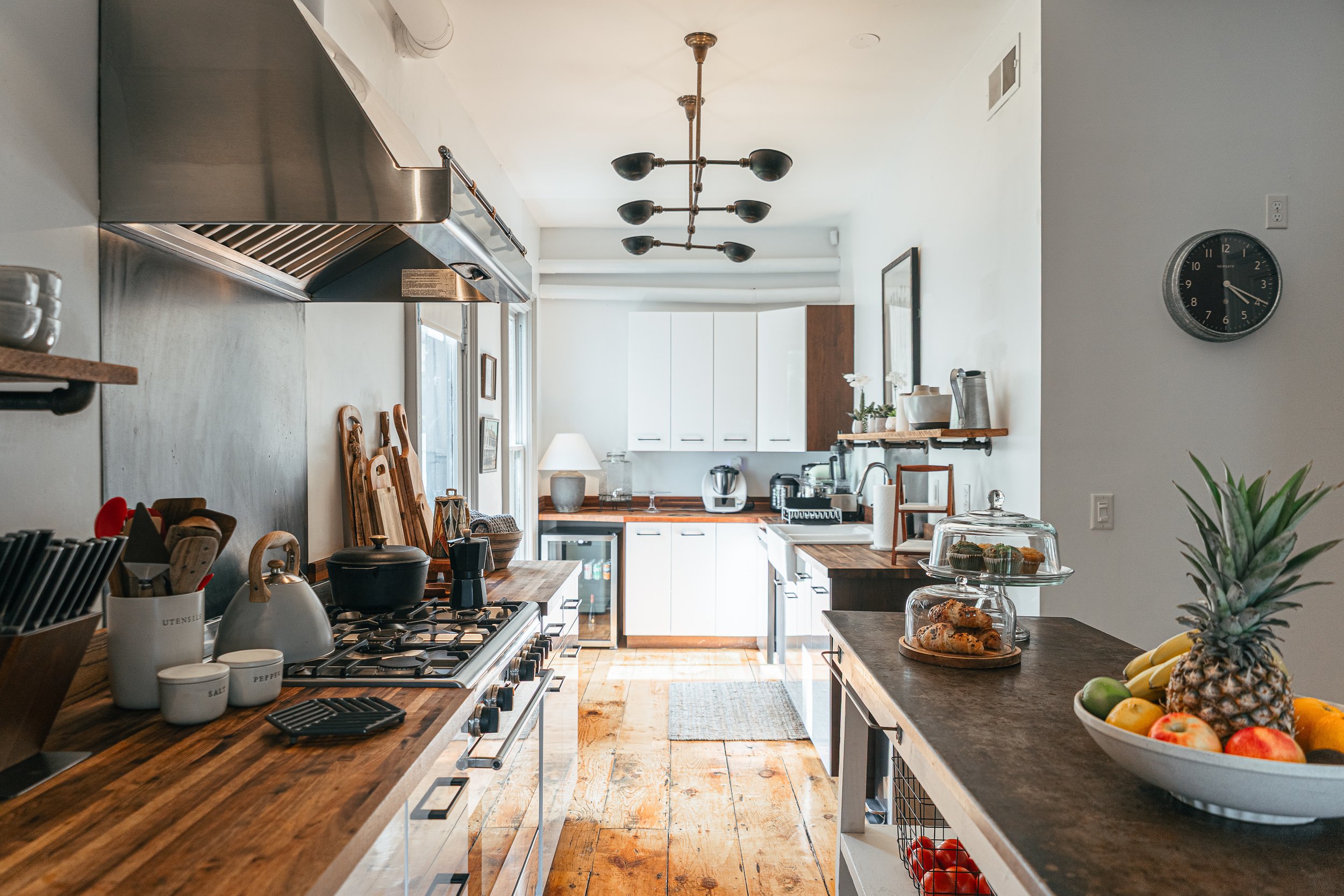Modern kitchen with wooden countertops, white cabinets, and stainless steel appliances. A bowl of tropical fruits including pineapple, bananas, and apples is on the counter. There are cake stands with muffins or cupcakes and a pineapple on a side table. Pendant lights hang from the ceiling, and a clock on the wall shows the time as 3:03.