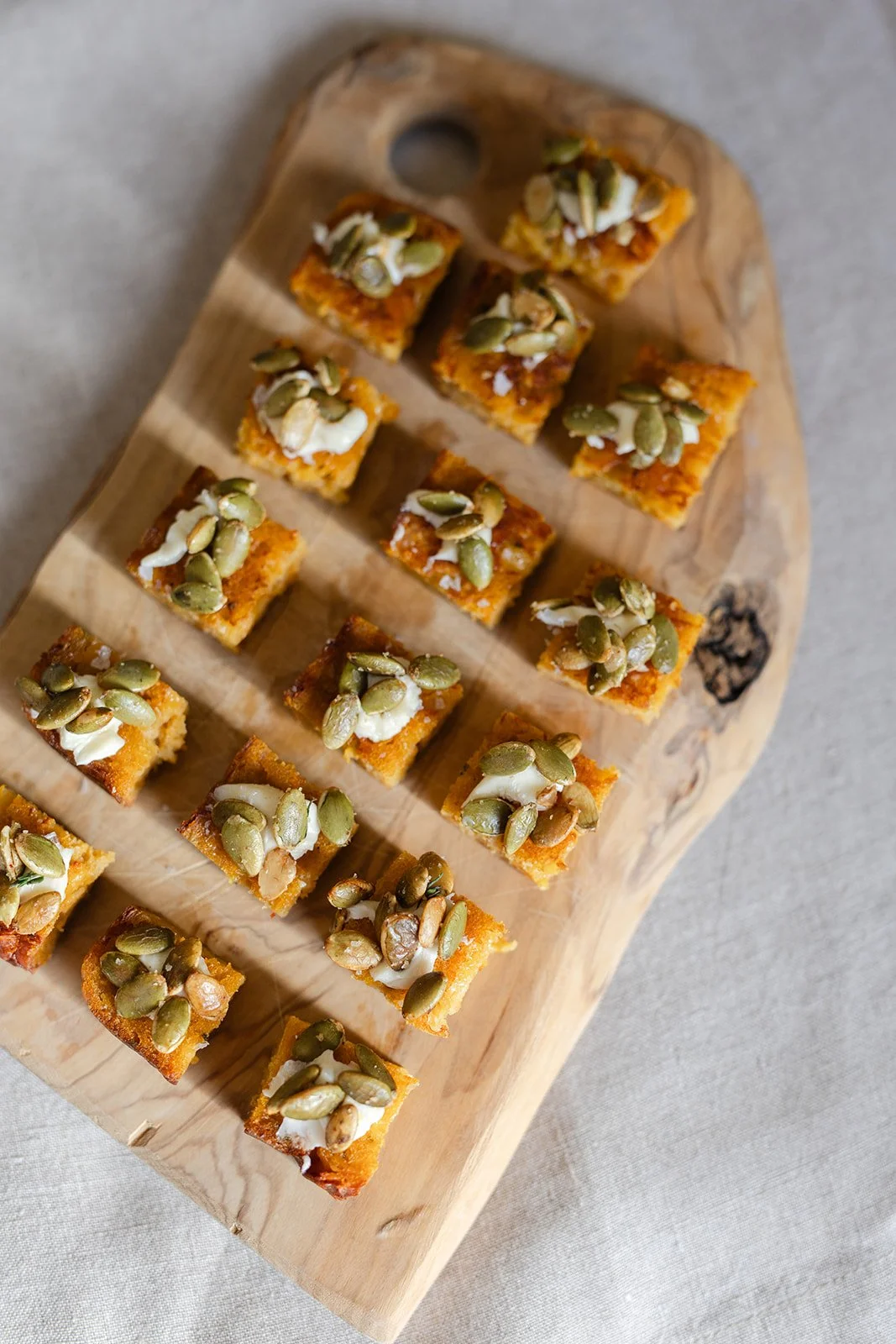Small square pieces of golden-brown toast topped with white sauce and green pumpkin seeds on a wooden serving board.