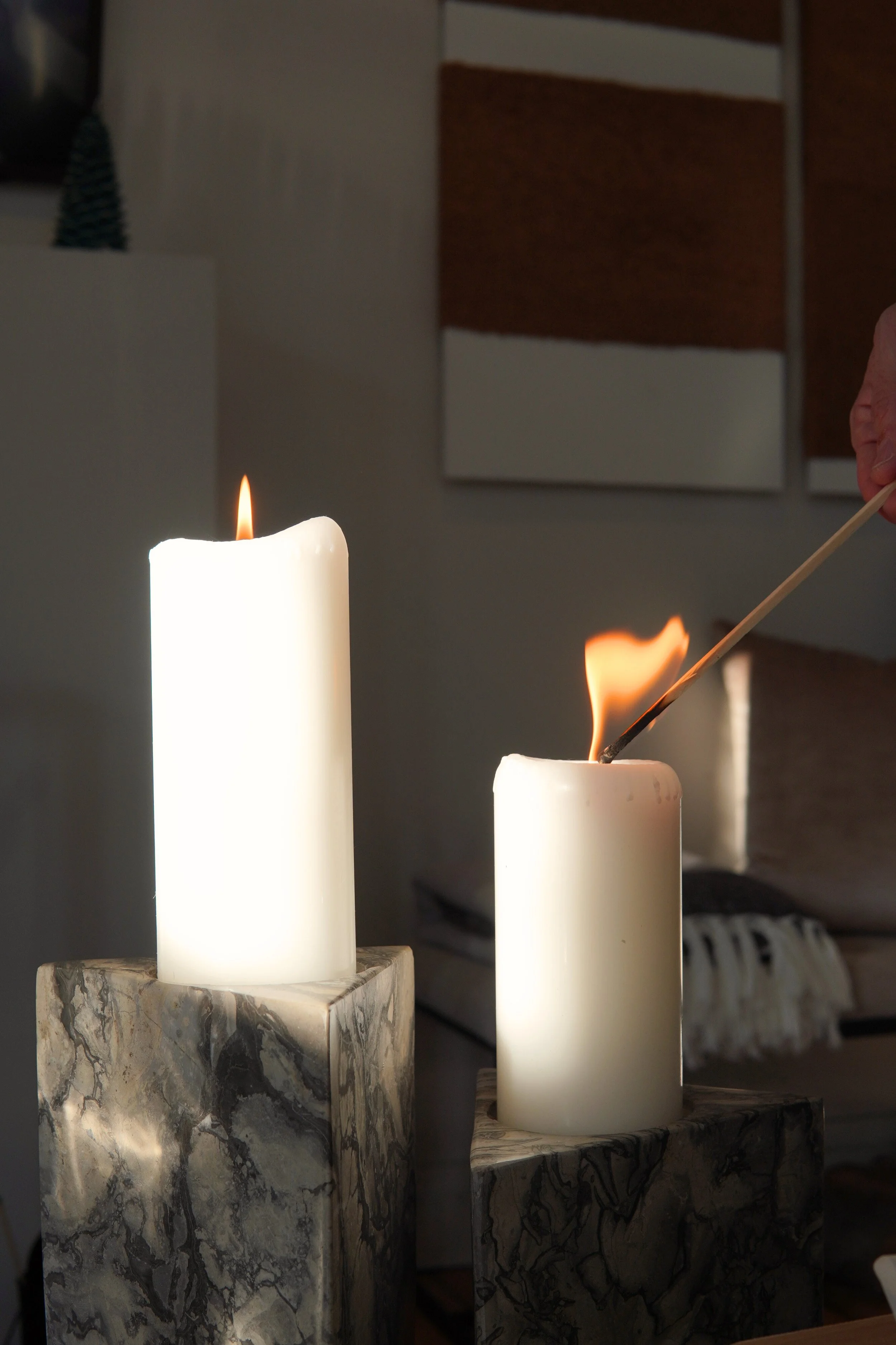 Two white candles placed on marble bases, with one being lit and having a flame, in a cozy indoor setting.