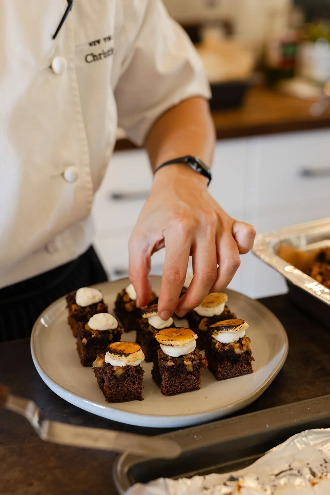 A chef in a white uniform garnishes chocolate dessert squares with toasted marshmallows and whipped cream on a plate.