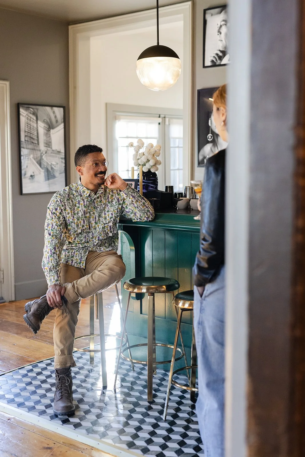 A man with glasses, a patterned shirt, and beige pants sitting on a barstool, smiling and talking to a woman standing nearby, in a cozy cafe or bar setting with a green bar counter and wall art.