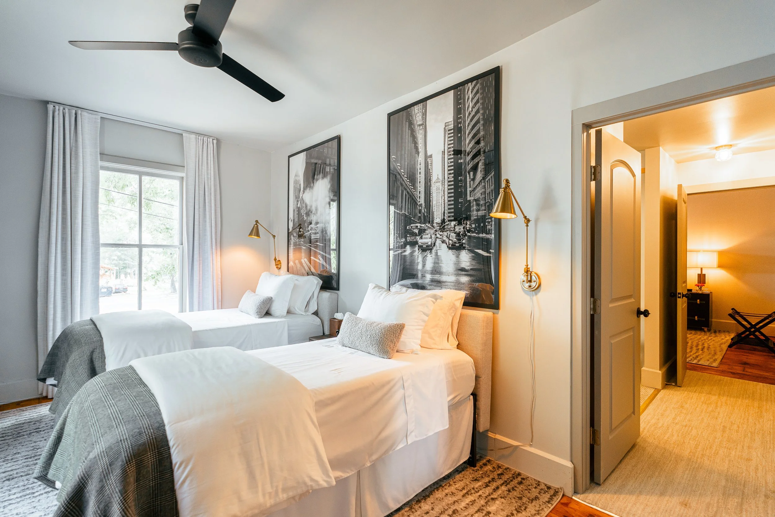 A bedroom with two twin beds with white bedding and plaid blankets, each with a white pillow and a gray decorative pillow. The room has a window with white curtains, black and white cityscape artwork on the walls, and gold reading lamps above each bed. There is a ceiling fan and a doorway leading to a lit hallway with a dresser and a suitcase rack.