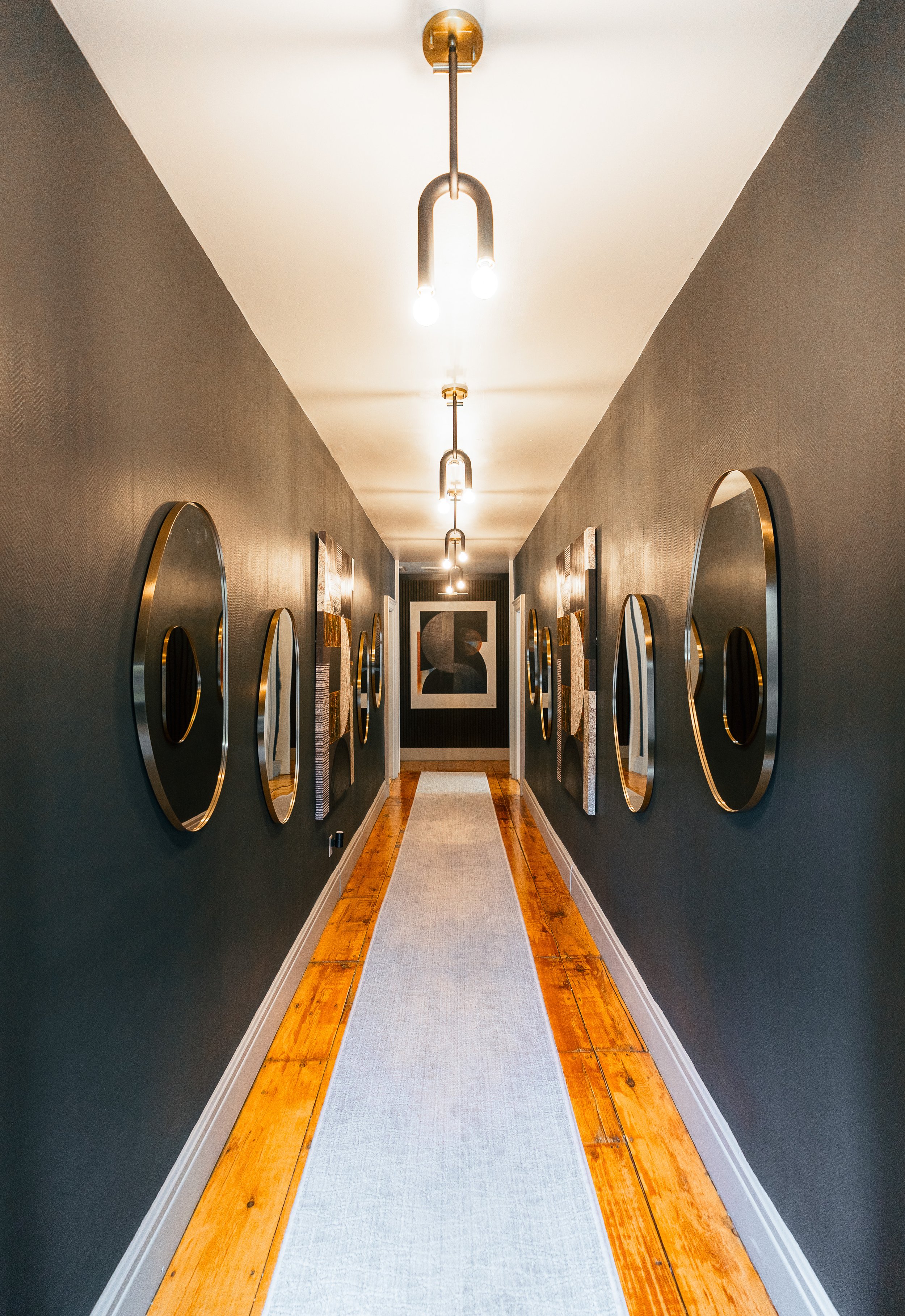 A narrow hallway with gray walls, decorated with modern oval mirrors with gold frames, hung on both sides. The corridor is lit by modern ceiling fixtures and has a light-colored runner rug on a wooden floor, leading to a framed art piece at the far end.