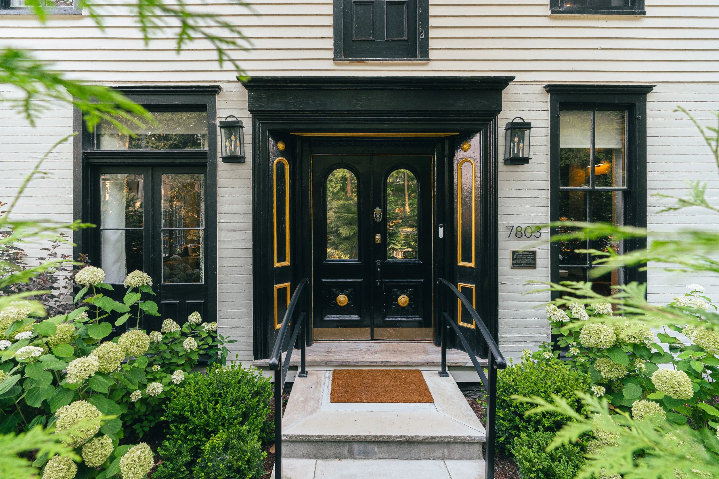 Front entrance of a house with black double doors, black and gold trim, black railings, surrounded by lush green bushes and white flowering plants, with house number 7803 visible.