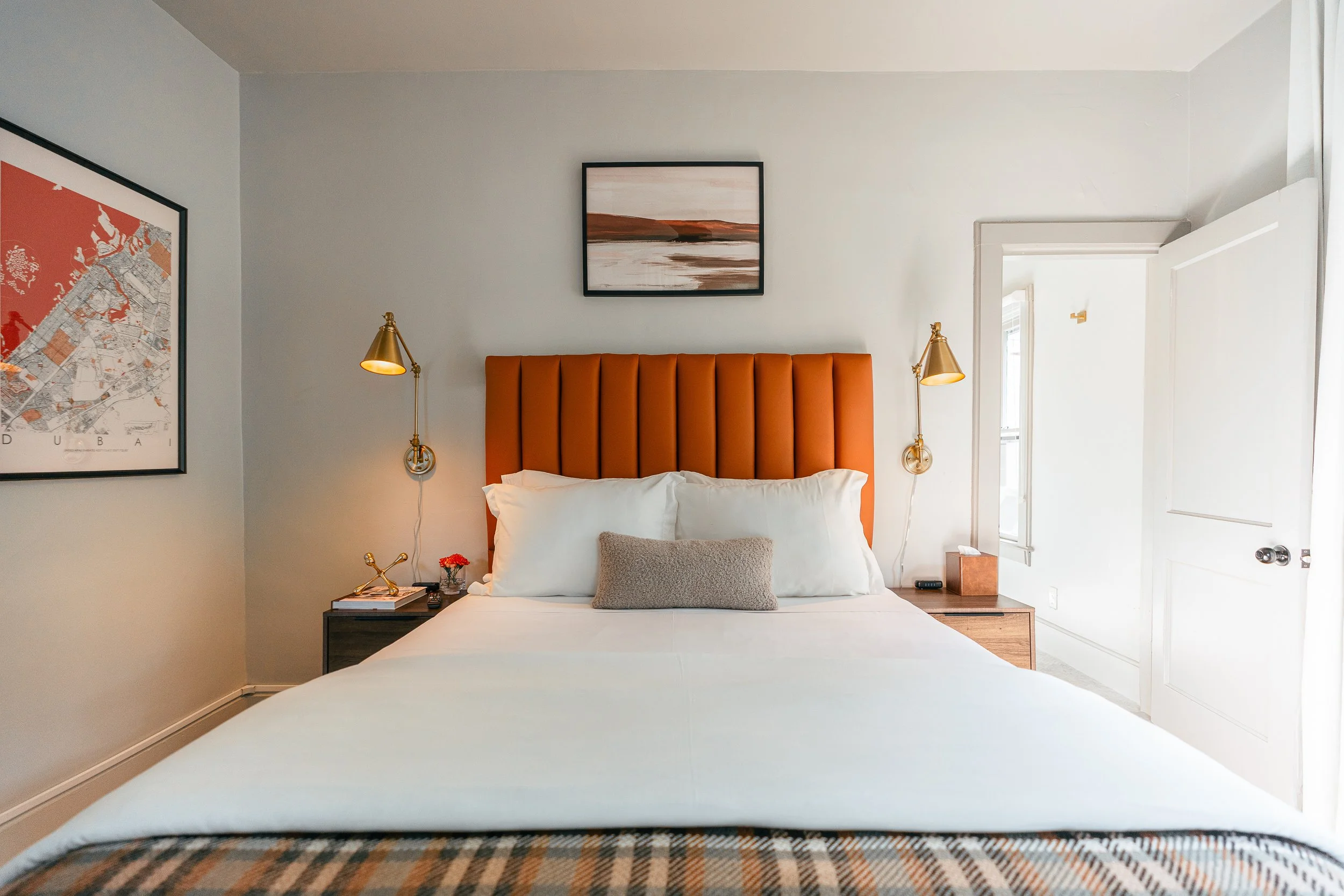 A neatly made bed with an orange upholstered headboard, white pillows, a gray bolster pillow, and a white duvet in a modern bedroom. Two gold wall-mounted reading lamps are on either side of the bed, with bedside tables holding books, a small flower, and a tissue box. There are framed pictures on the wall, and a partially open white door leads to another room with natural light.