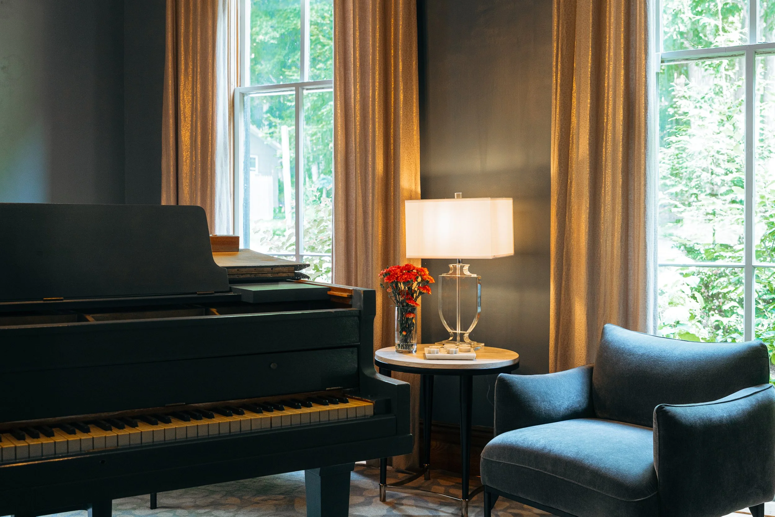 A cozy living room corner with a black upright piano, a round side table with a white lamp and a vase of red flowers, and a blue armchair. Two large windows with beige curtains let in natural light.