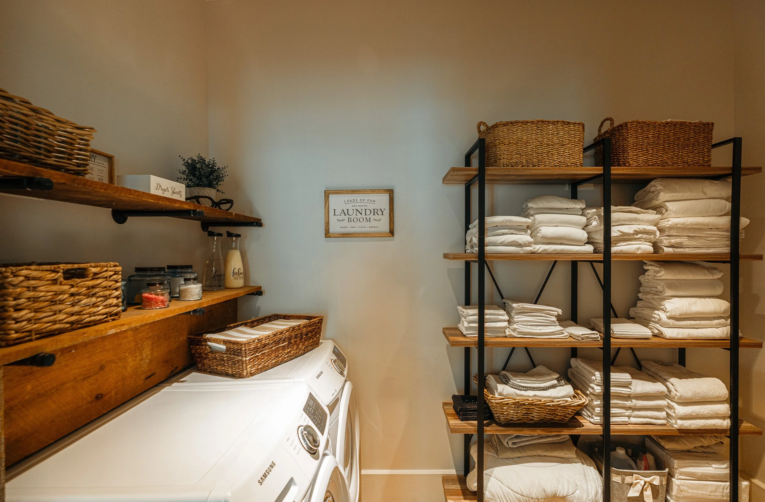 A laundry room with wooden shelves holding baskets, folded towels, and linens, and a washing machine below the shelves.
