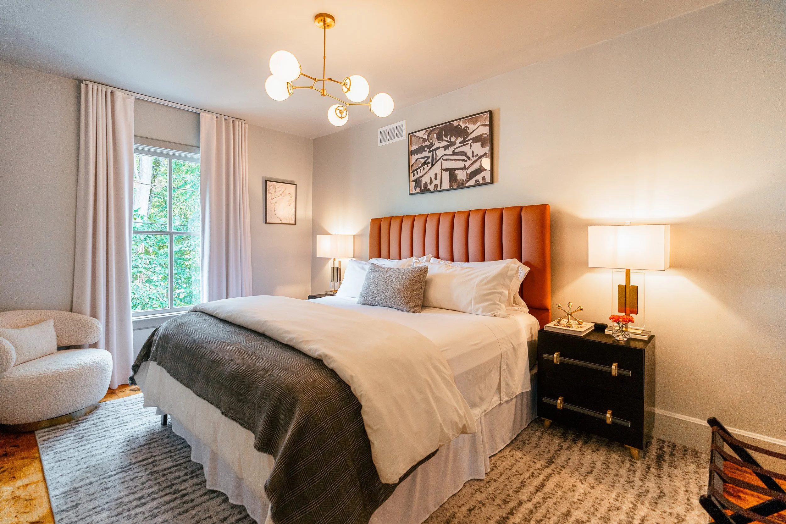 A bedroom with a large bed, orange headboard, white bedding, and decorative pillows. There are two nightstands with lamps, a window with white curtains, a beige armchair, and wall art. A ceiling light fixture is also visible.