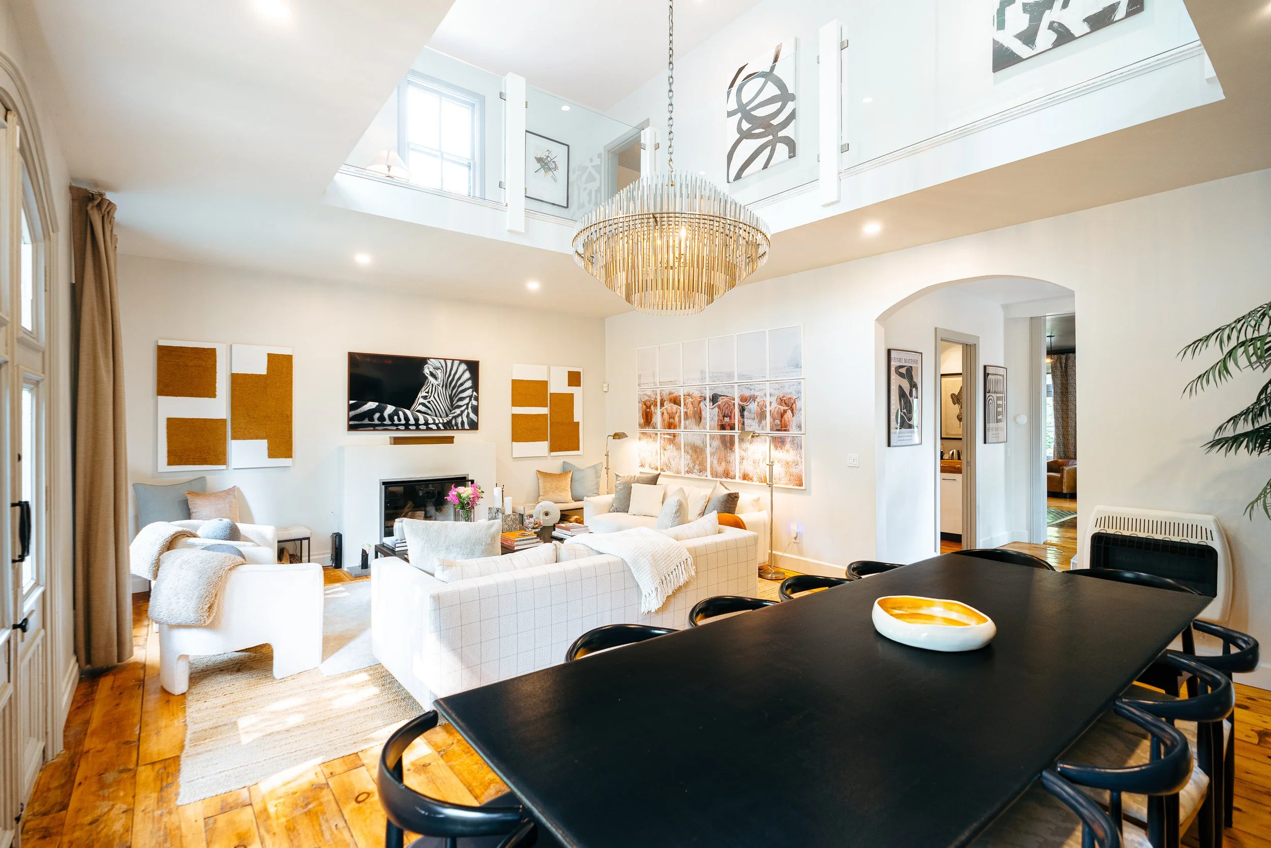 Bright living room with white sofas, artwork on walls, a chandelier, a flat-screen TV above a fireplace, a large black dining table with black chairs, and decorative plants.