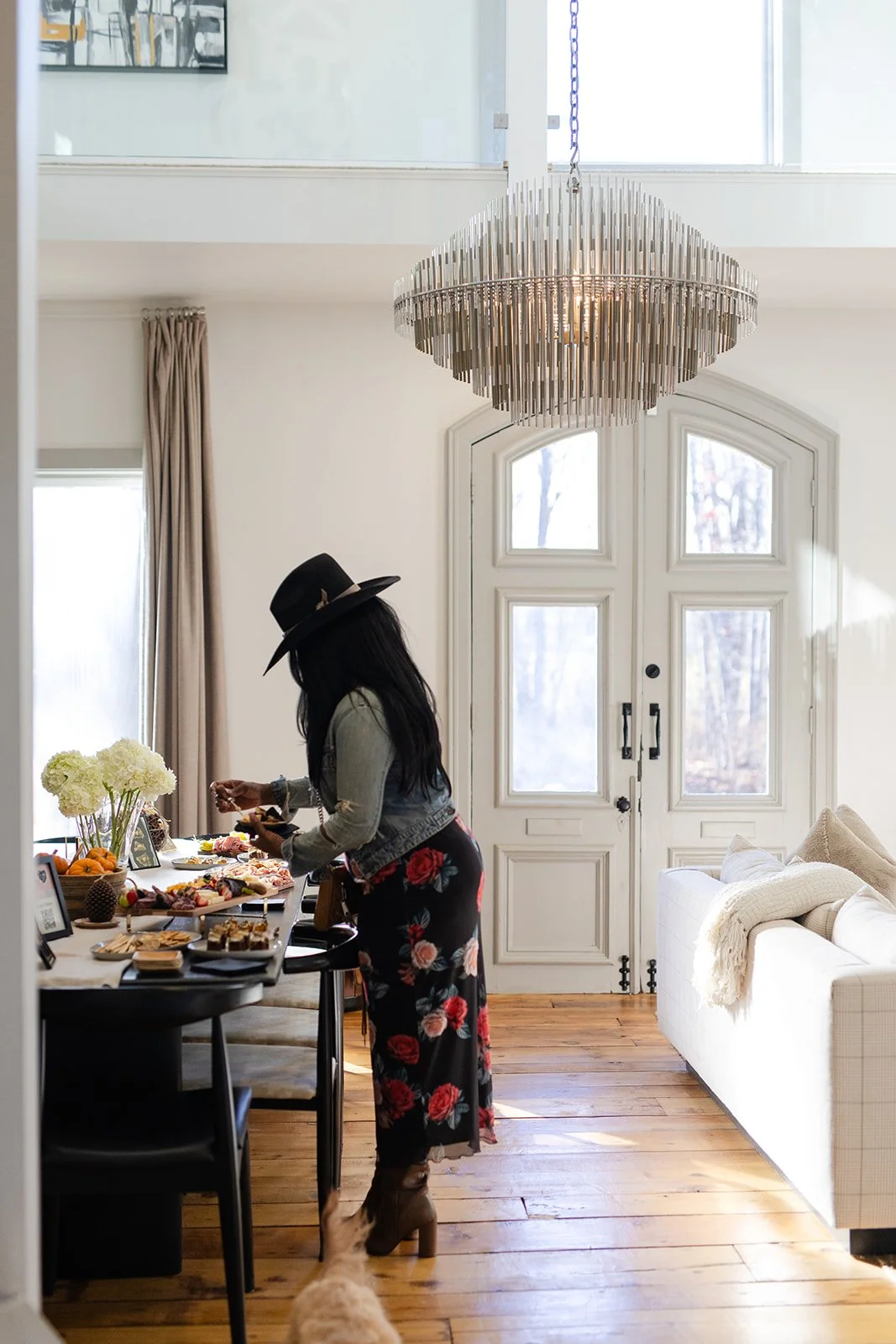 Woman with long dark hair, wearing a floral skirt, gray jacket, and a black wide-brimmed hat, preparing food at a dining table in a bright, modern room.