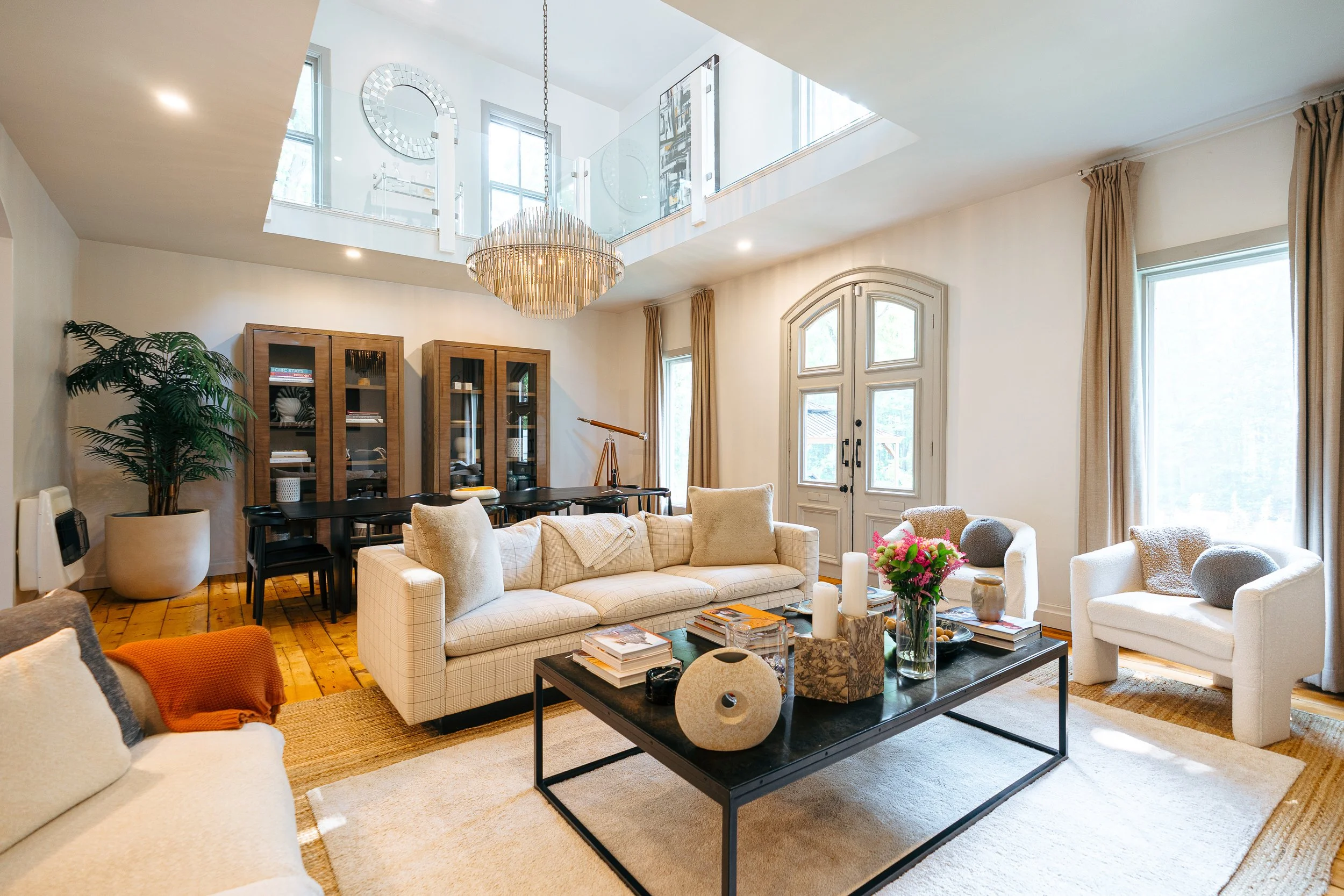 A spacious and well-lit living room with white walls, hardwood floors, and large windows. It features modern furniture including a beige sofa with pillows, a white armchair, a black coffee table with decorative items and books, a dining table with chairs, wooden display cabinets, a large plant, and a chandelier hanging from the ceiling.