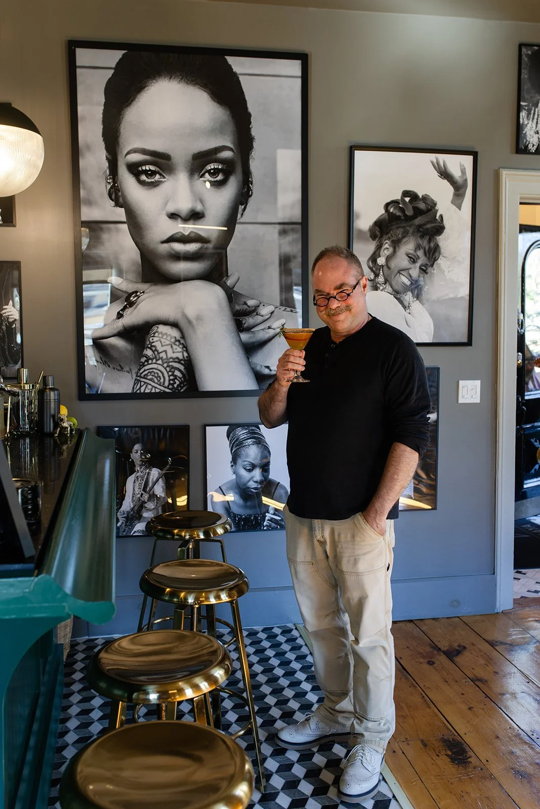 A man with glasses holding a cocktail in a bar with black and white portraits of women on the wall behind him.