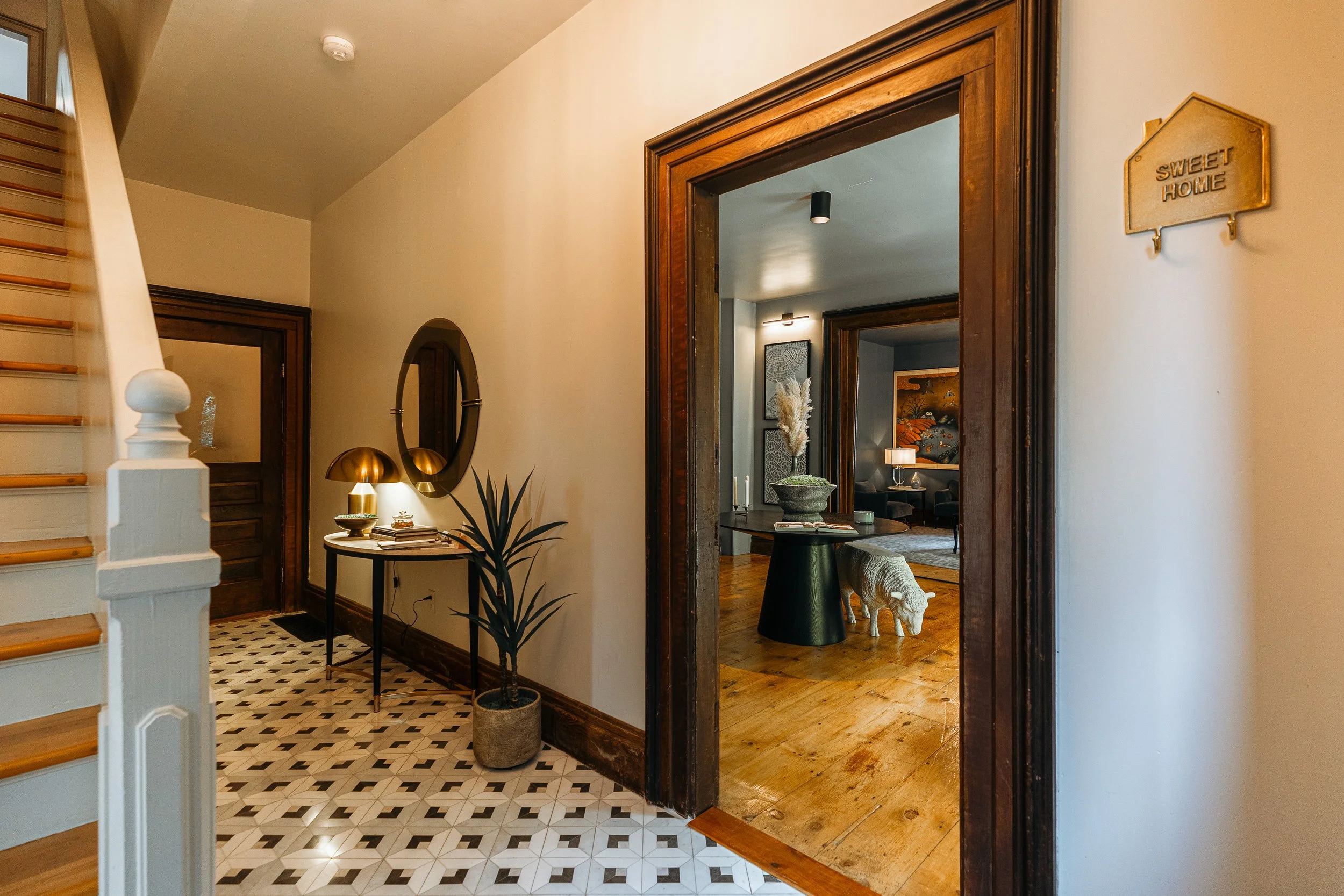 View of an entryway with a staircase on the left, a round mirror and table with lamps and books on the right, a potted plant, and open doorways leading to a living room with wooden flooring and decorative art.