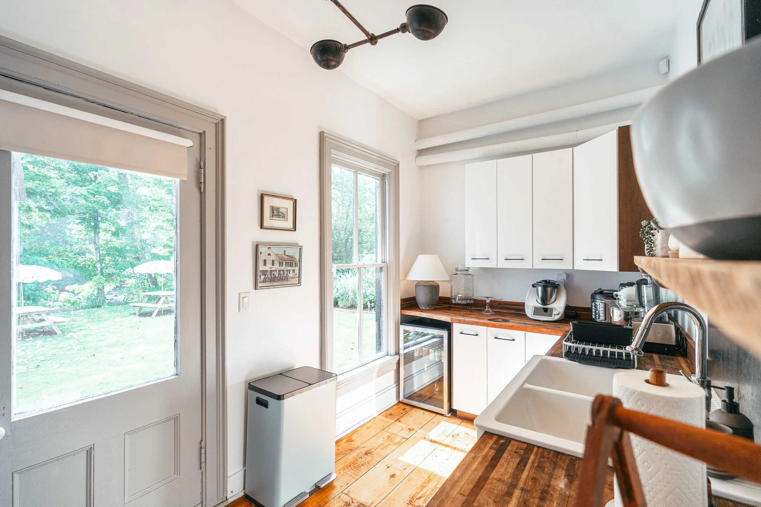 Small kitchen area with white cabinets, wooden countertop, a window, countertop appliances, and a door leading outside to a grassy patio with picnic tables