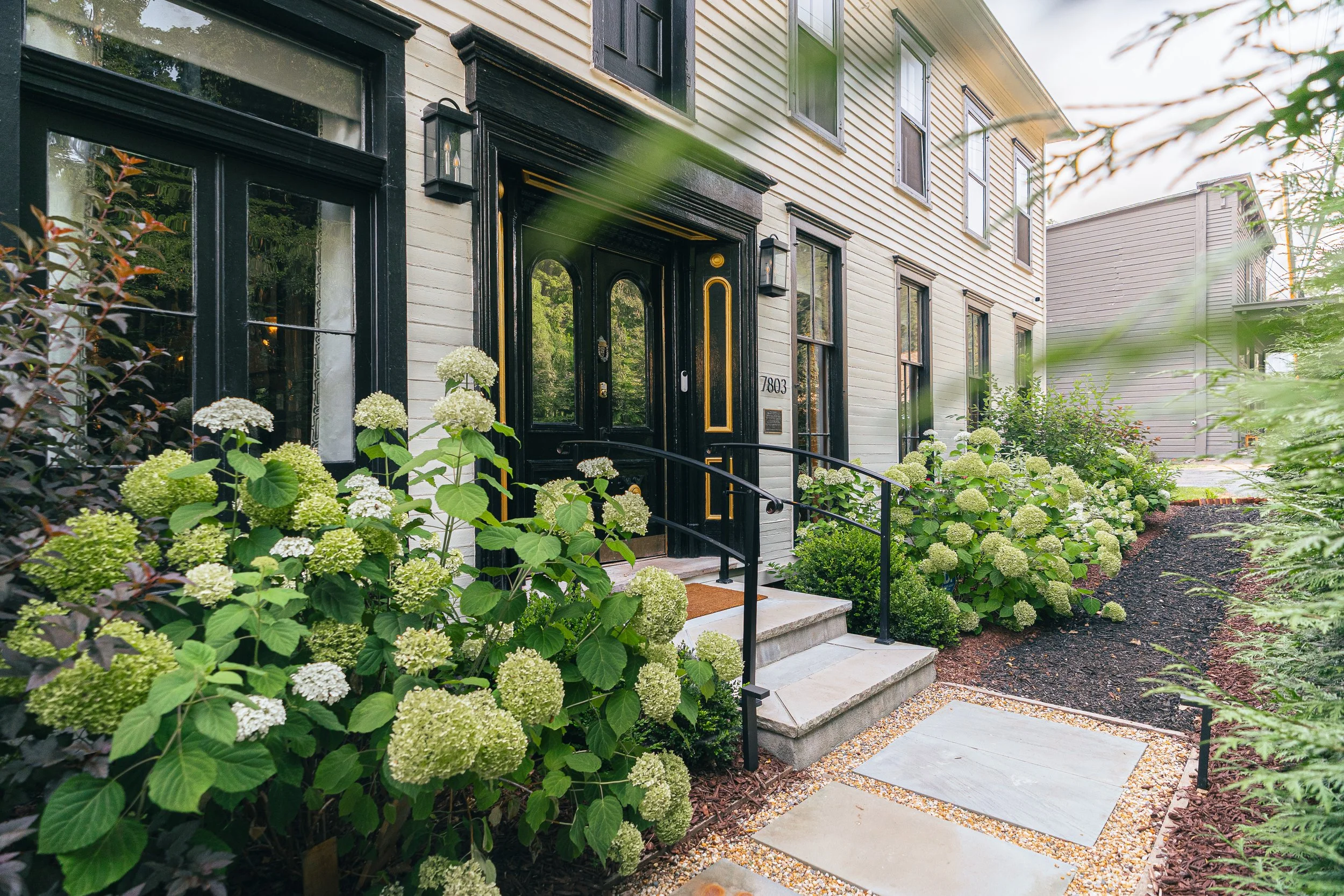 Entrance of a house with black door and golden accents, surrounded by lush green bushes and white flowering plants, with stone steps and a pathway leading to it.