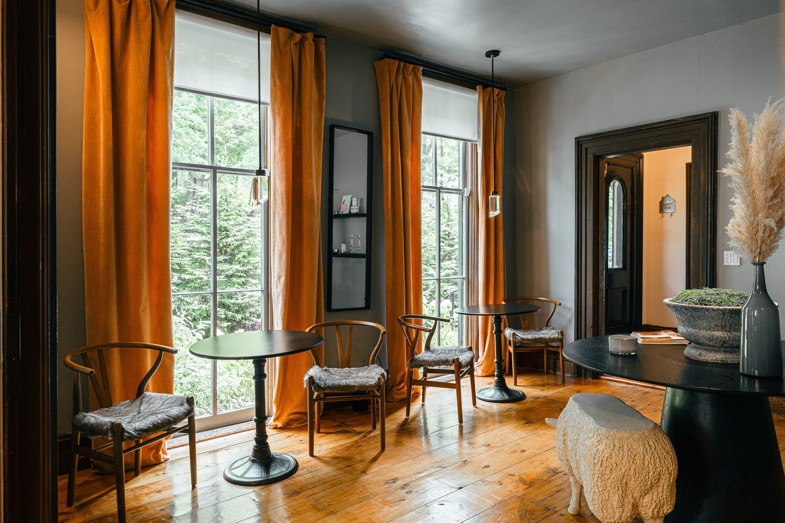 Interior of a room with wooden floors, large windows with orange curtains, and a black door. There are three small round black tables with chairs that have furry cushions, and a black table with a large stone planter and decorative items.