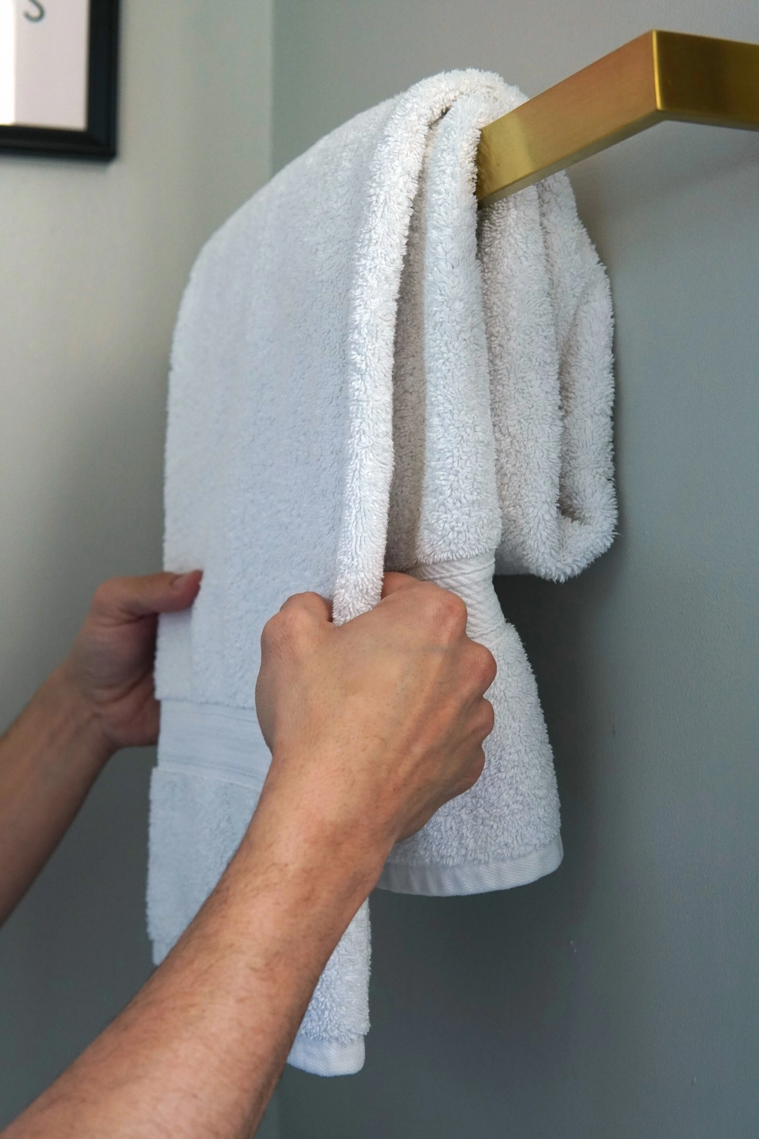 Person hanging a white towel on a gold-colored towel bar on a light gray wall.