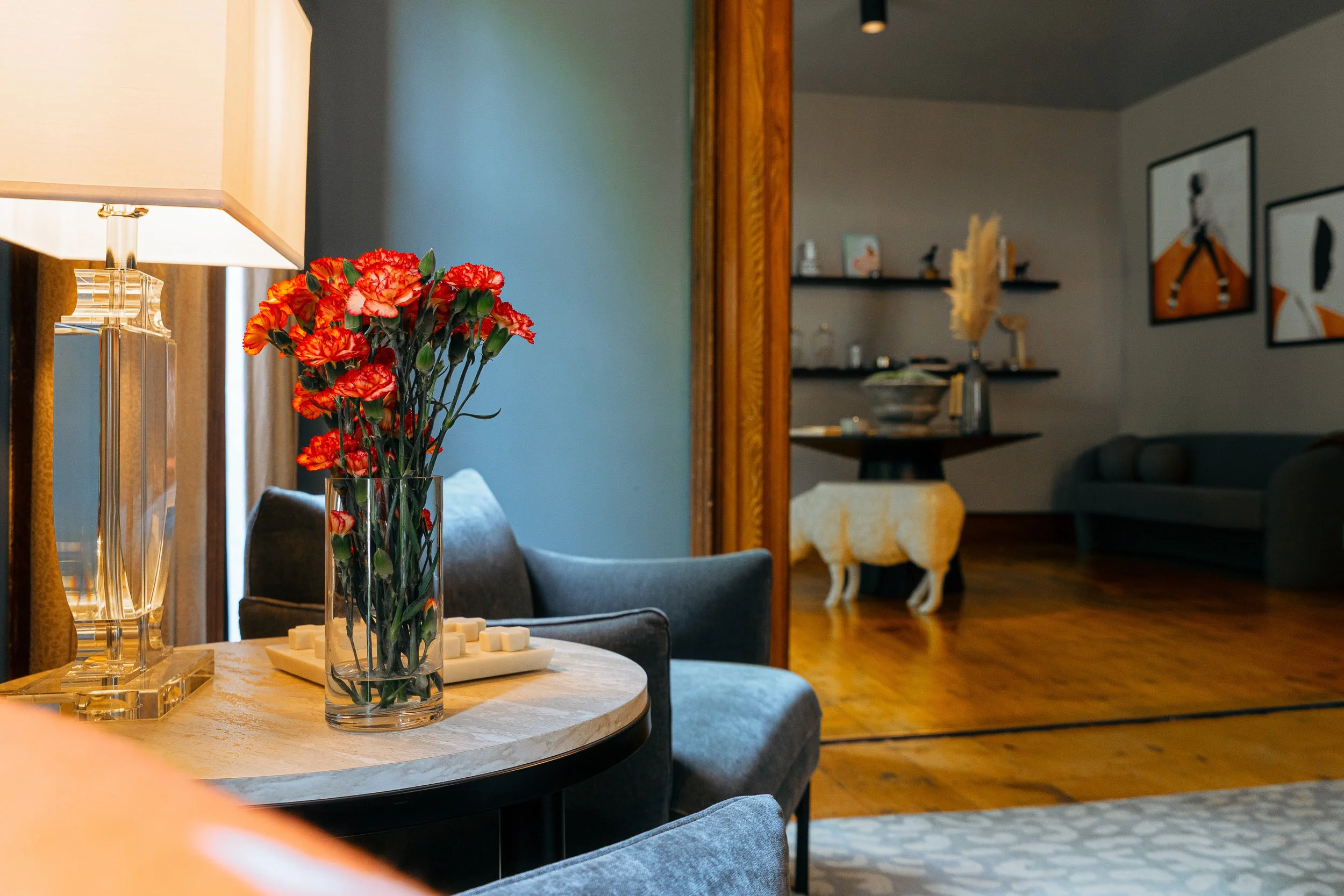 A vase with red and orange flowers on a marble side table in a living room, with a lamp beside it and a gray armchair. In the background, a room with wood flooring, abstract wall art, a gray sofa, and decorative items on black shelves.