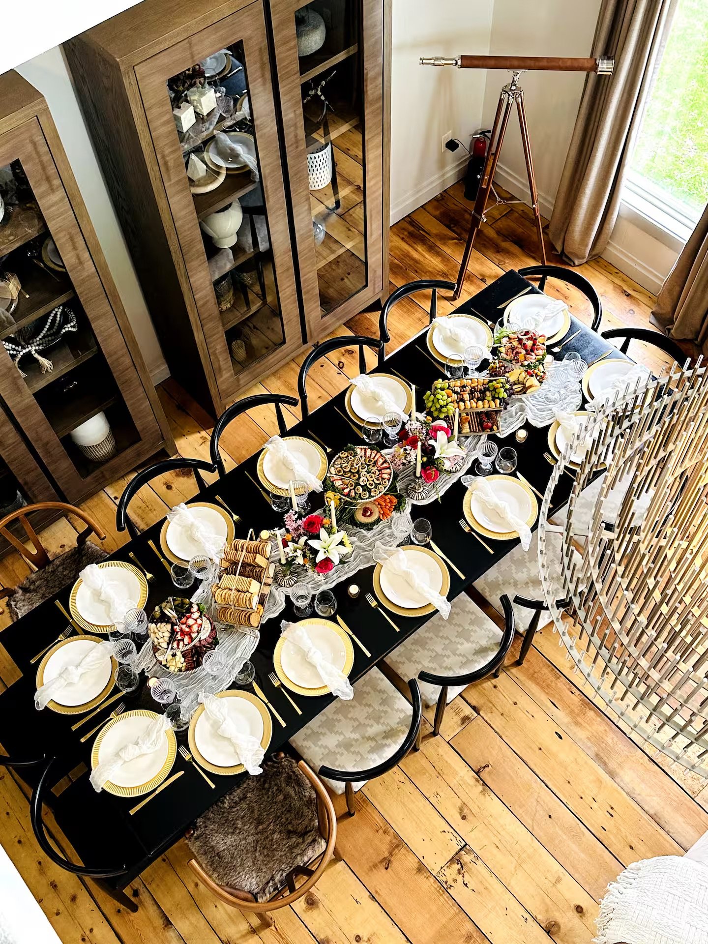 Overhead view of a dining table set for a celebration, with plates, napkins, glassware, and decorative centerpieces, adjacent to wooden china cabinets and a telescope near a window.