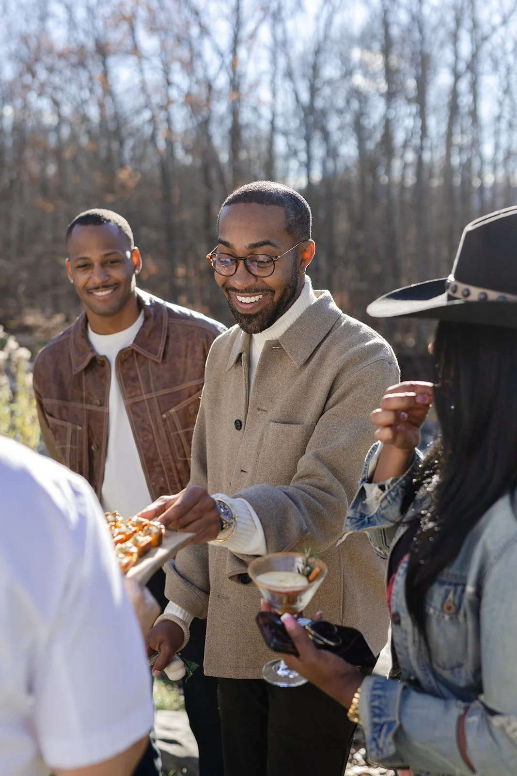 Group of four friends outdoors, smiling and enjoying snacks and drinks during daytime in a wooded area.