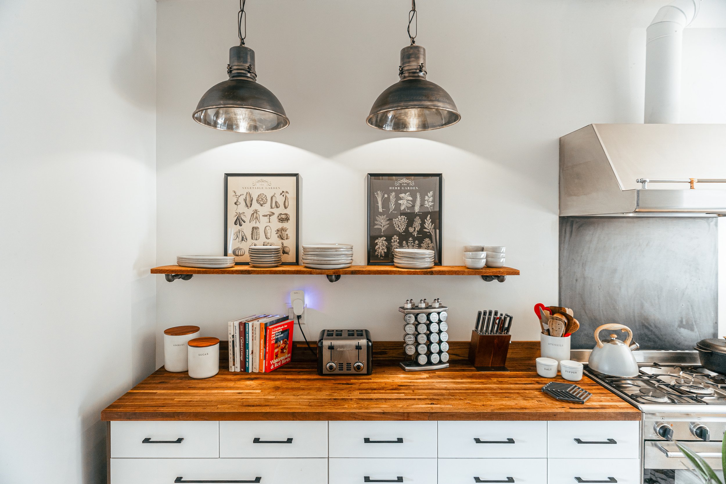 Kitchen with wooden countertop, white cabinets, shelves with dishes, framed vegetable garden and herb garden prints, toaster, spice rack, utensils, kettle, and stove with a gray backsplash, two metal pendant lights hanging above.