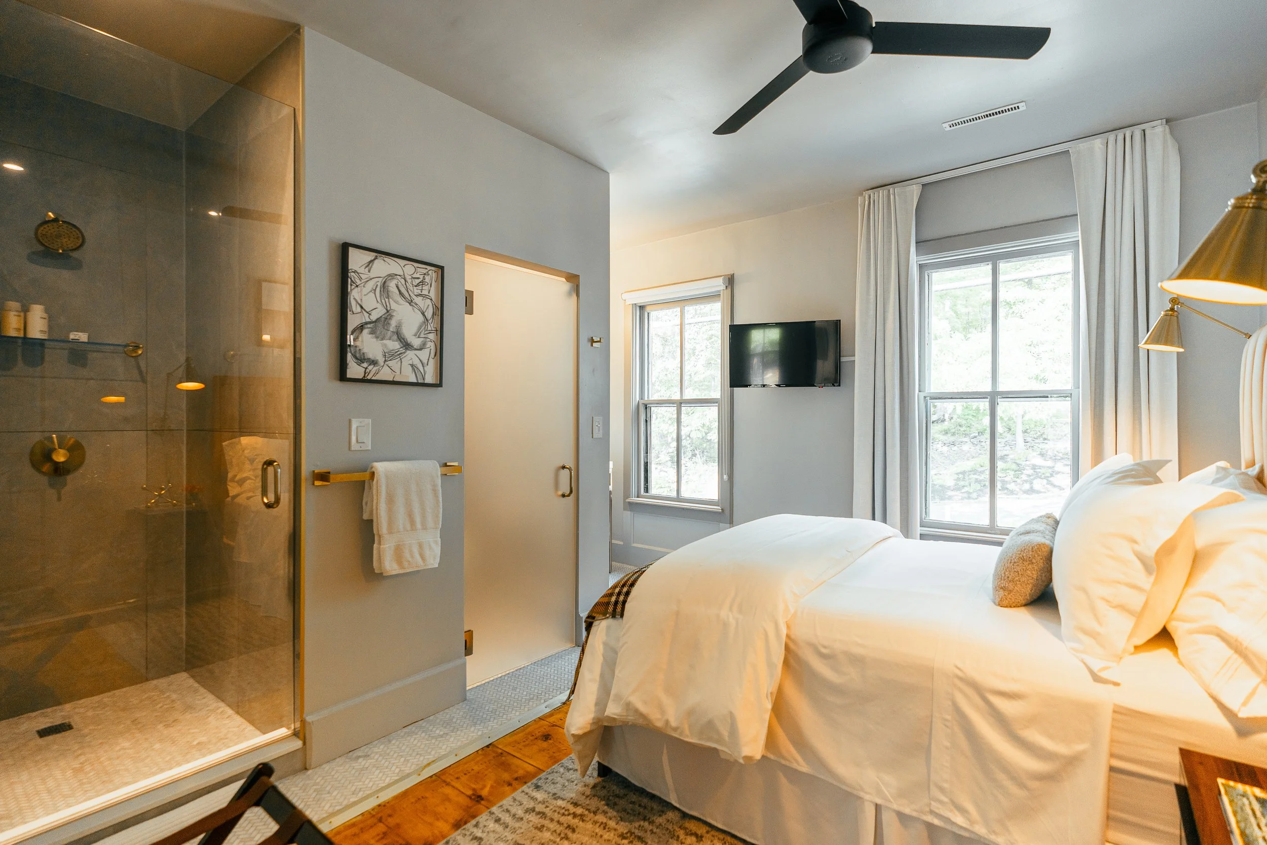 A bedroom with a bed, two windows, a wall-mounted TV, and a bathroom with a glass shower enclosure.