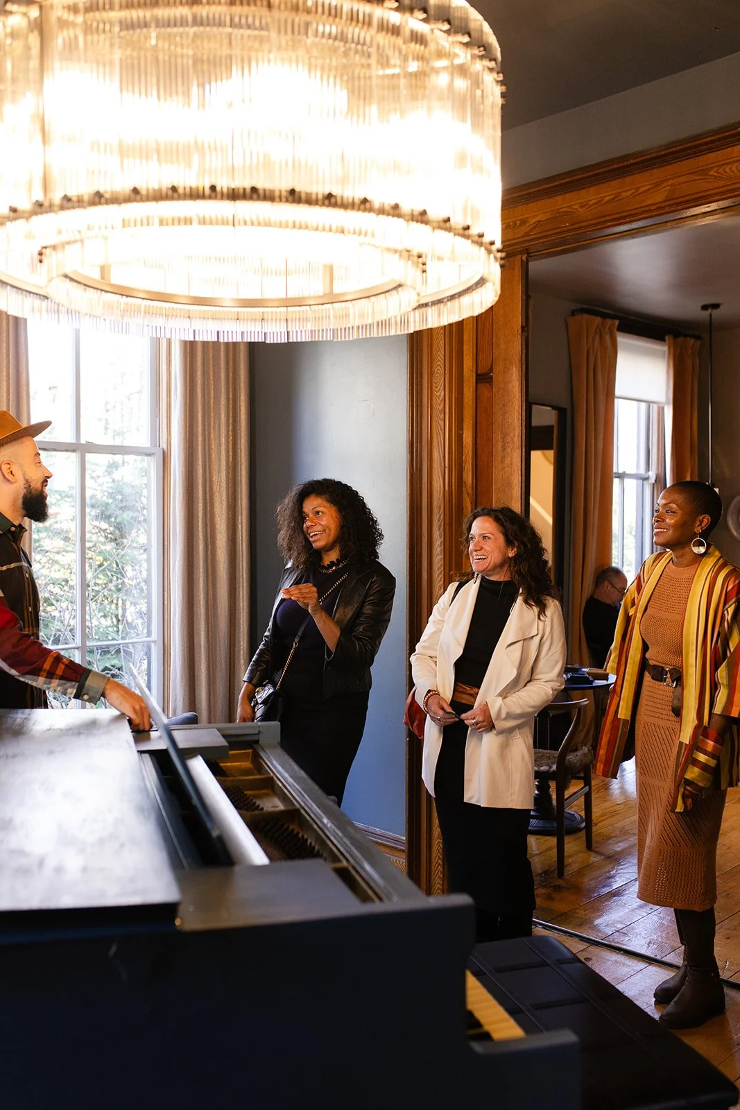A man plays the piano while four women watch and smile inside a warmly lit room with large windows and wooden trim.