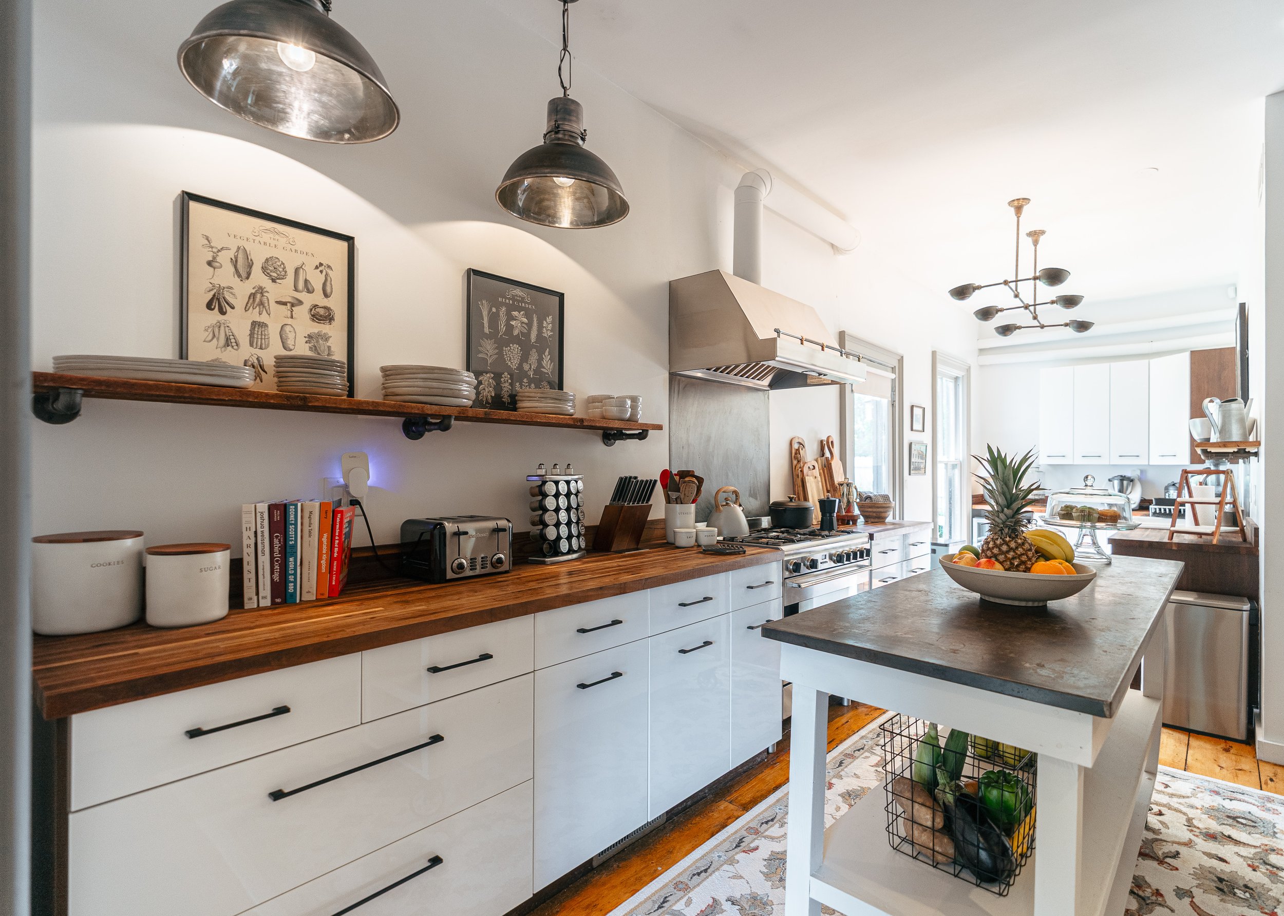 Modern kitchen with white cabinets and a wooden countertop. There are two framed vegetable illustrations on the wall, open shelves with plates and bowls, stainless steel appliances, and a kitchen island with a fruit bowl. The space features hanging ceiling lights, hardwood floors, and natural light.