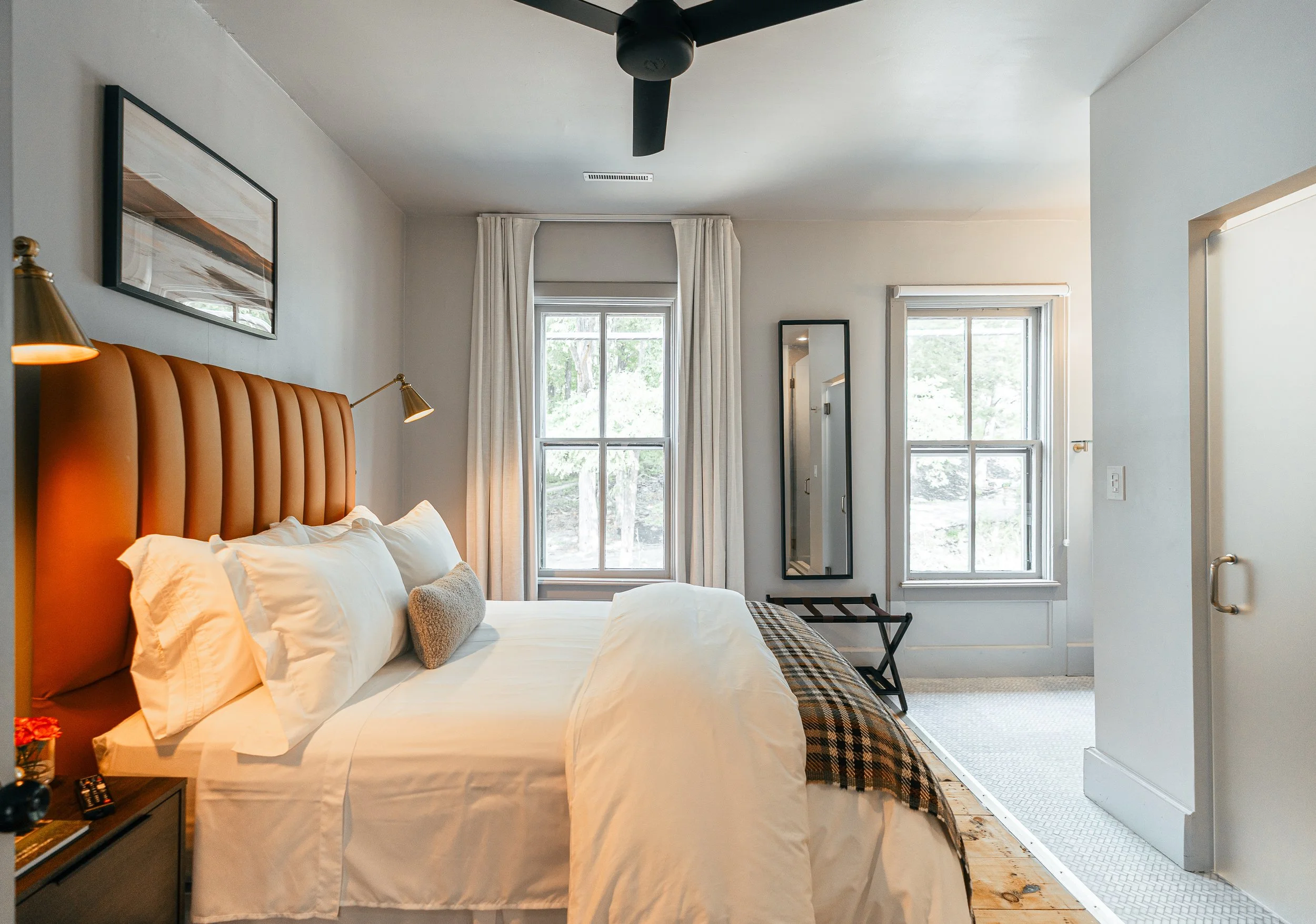 A cozy bedroom with a large bed, orange headboard, white bedding, and multiple pillows. Two large windows with white curtains let in natural light. There's a black framed mirror, a small bench, and a framed artwork on the wall. The room has a ceiling fan and a wooden floor with a runner rug.