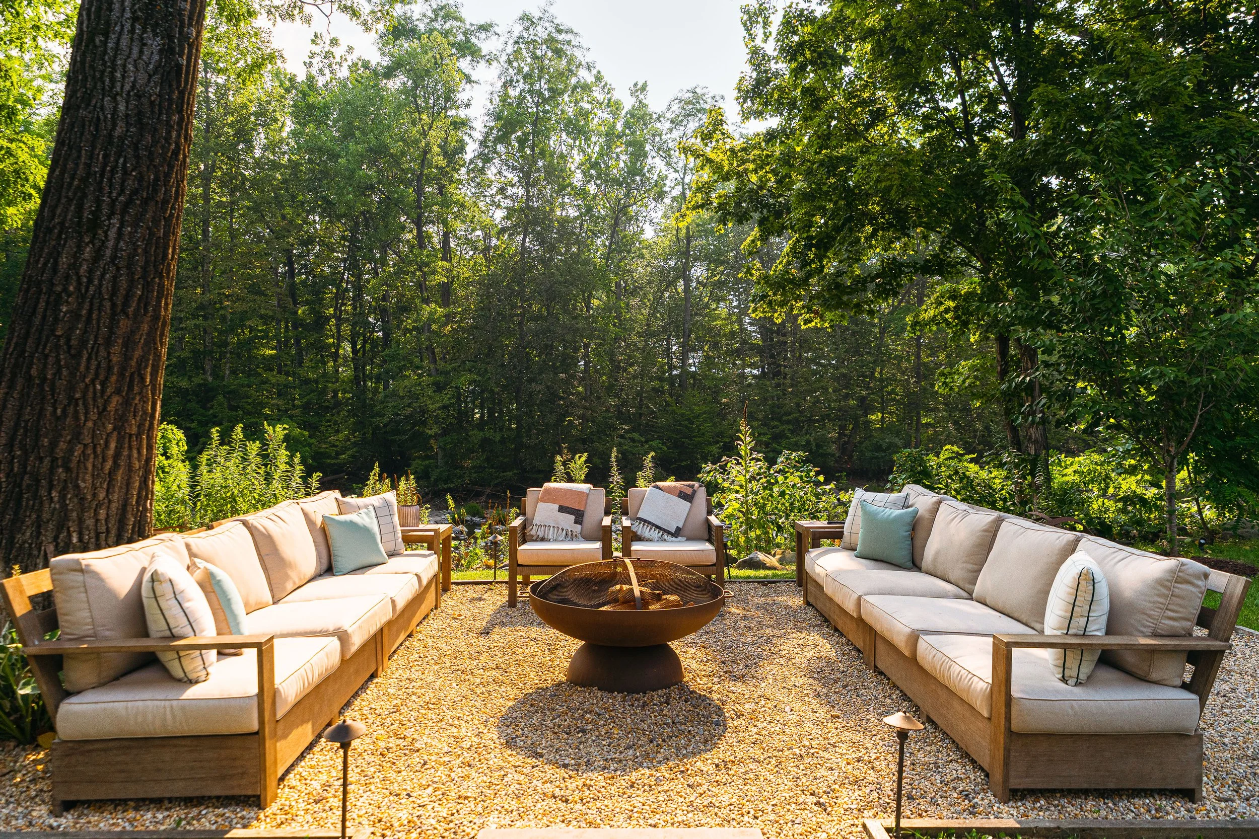 Outdoor patio area with beige sofas and chairs, a fire bowl in the center, surrounded by trees and green foliage