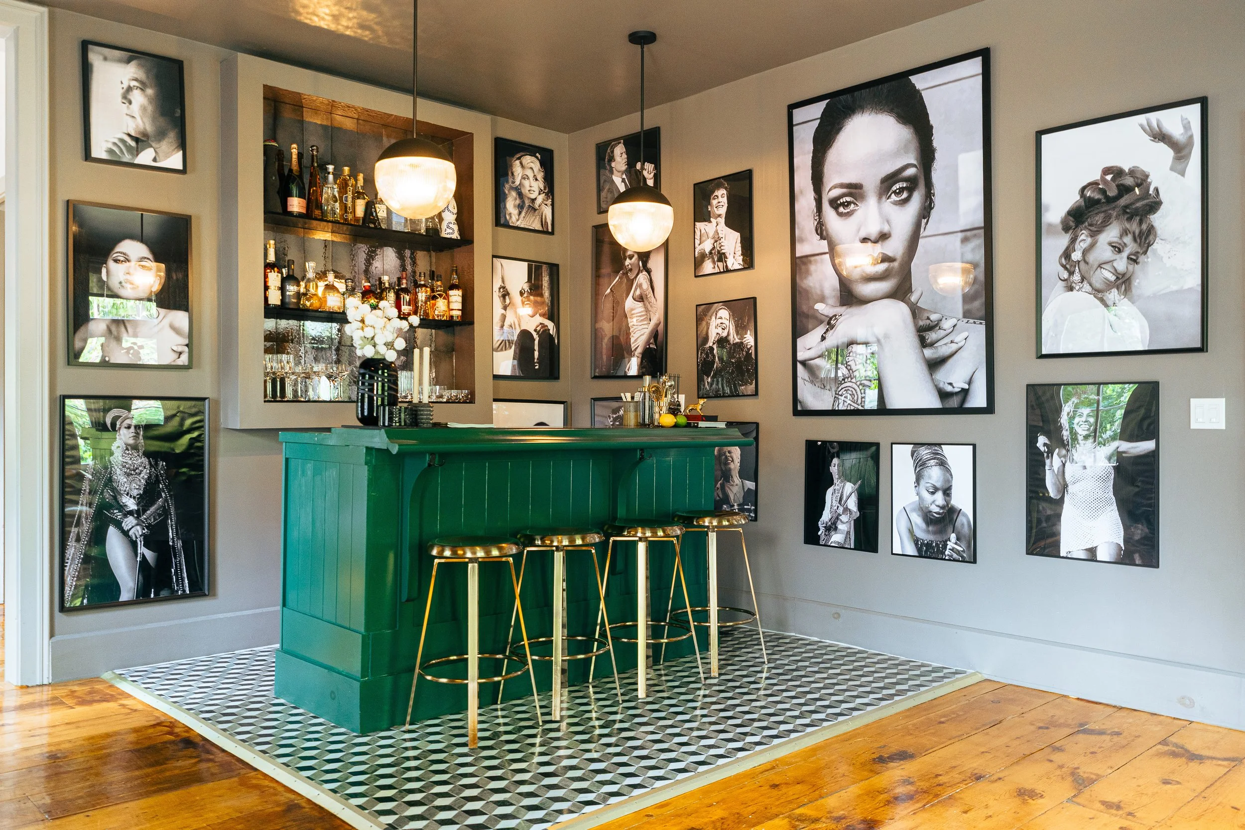 Interior of a bar with a green bar counter, gold bar stools, black and white portraits of women on the walls, and a shelf with liquor bottles and decorative items.