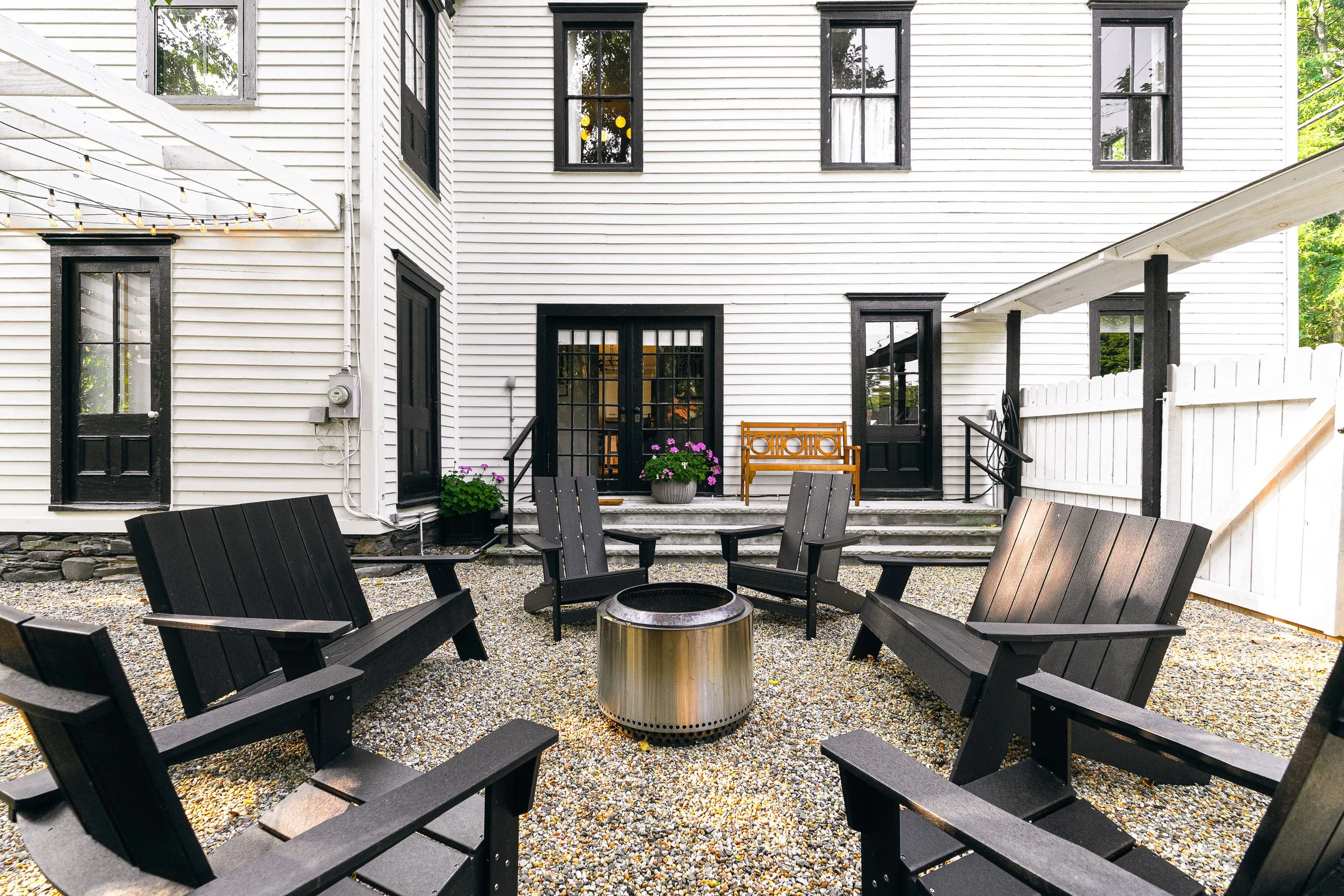 Outdoor patio with black Adirondack chairs arranged in a circle around a fire pit, gravel ground, white house with black framed windows, black doors, and flower pots with purple flowers on steps.