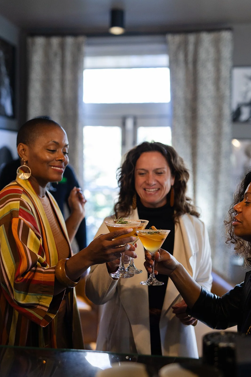 Three women in a social setting clinking glasses with cocktails, smiling, in a warmly lit room with large windows and curtains.