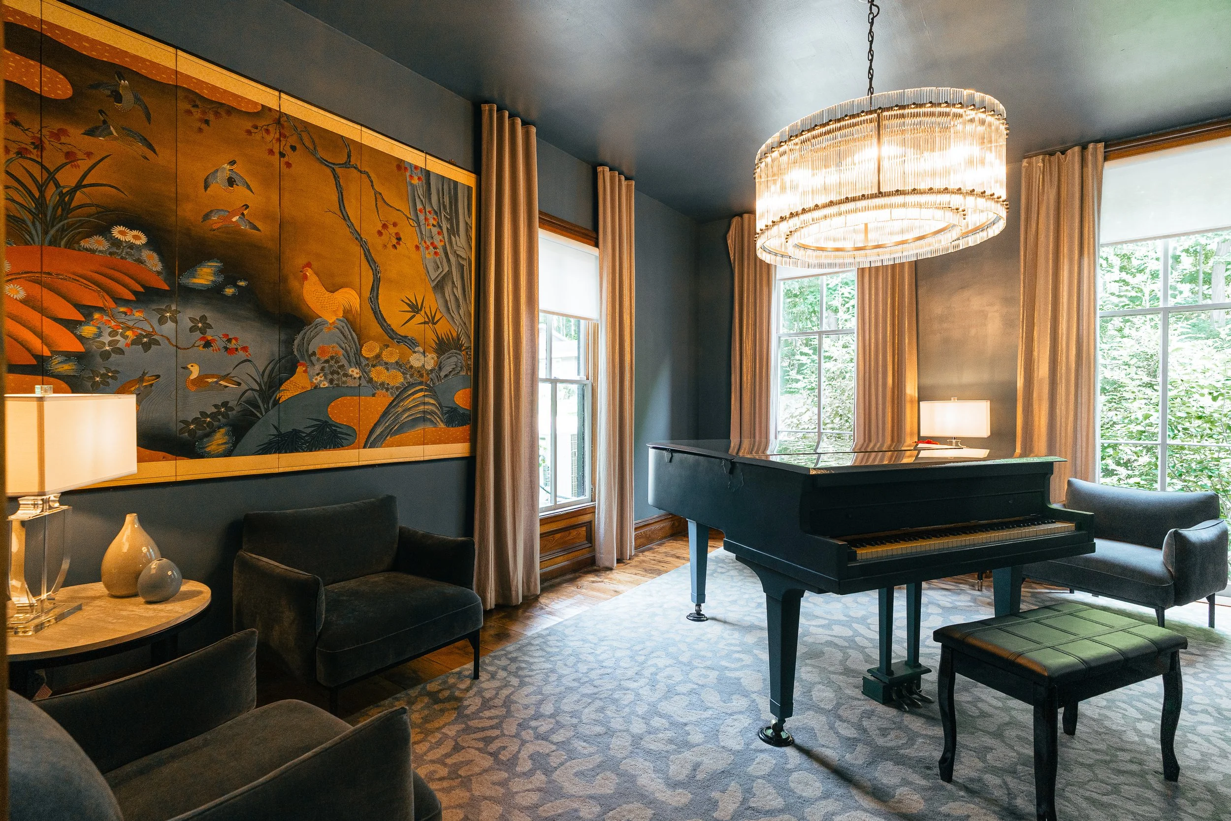 Elegant living room with a black grand piano, large chandelier, framed Asian art, and windows with beige curtains, brightening the space.